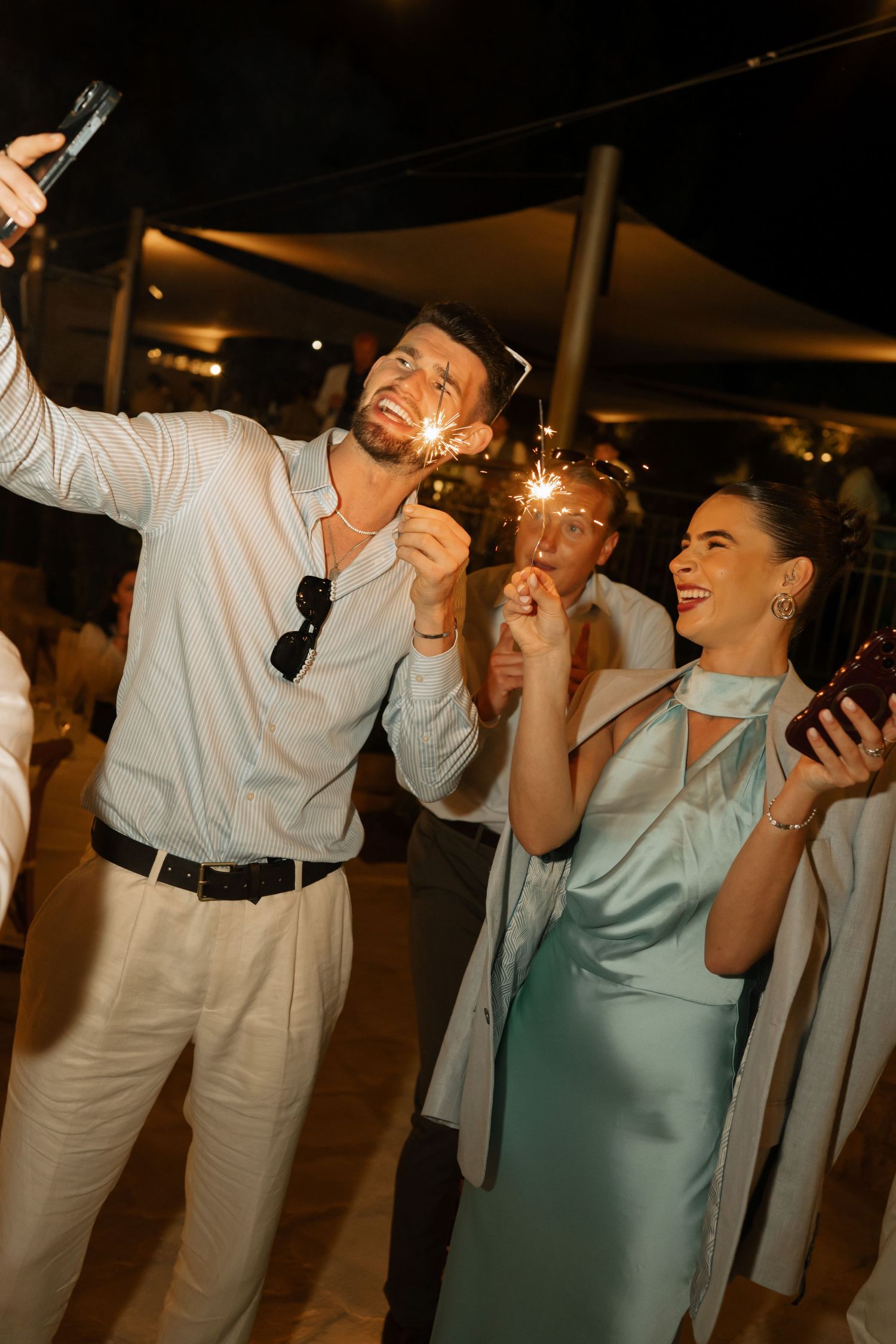 Couple laughing and holding sparklers at an outdoor evening celebration, dressed in elegant white and teal attire.