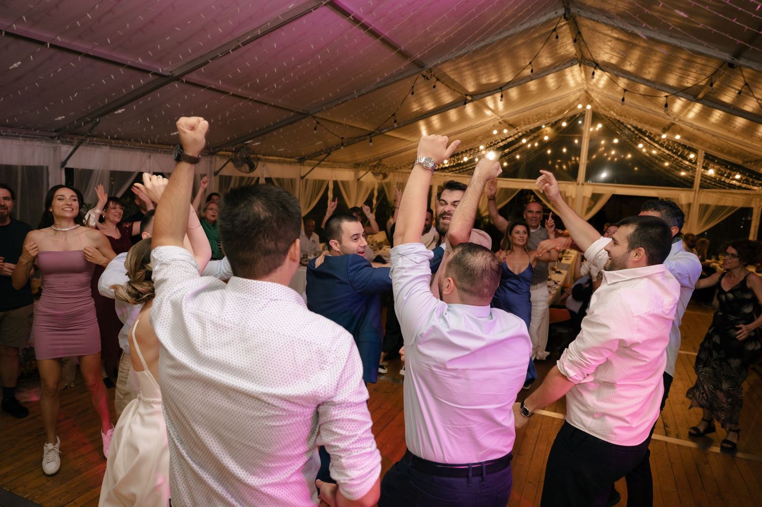Energetic wedding party dancing and celebrating on a wooden dance floor under warm lighting.