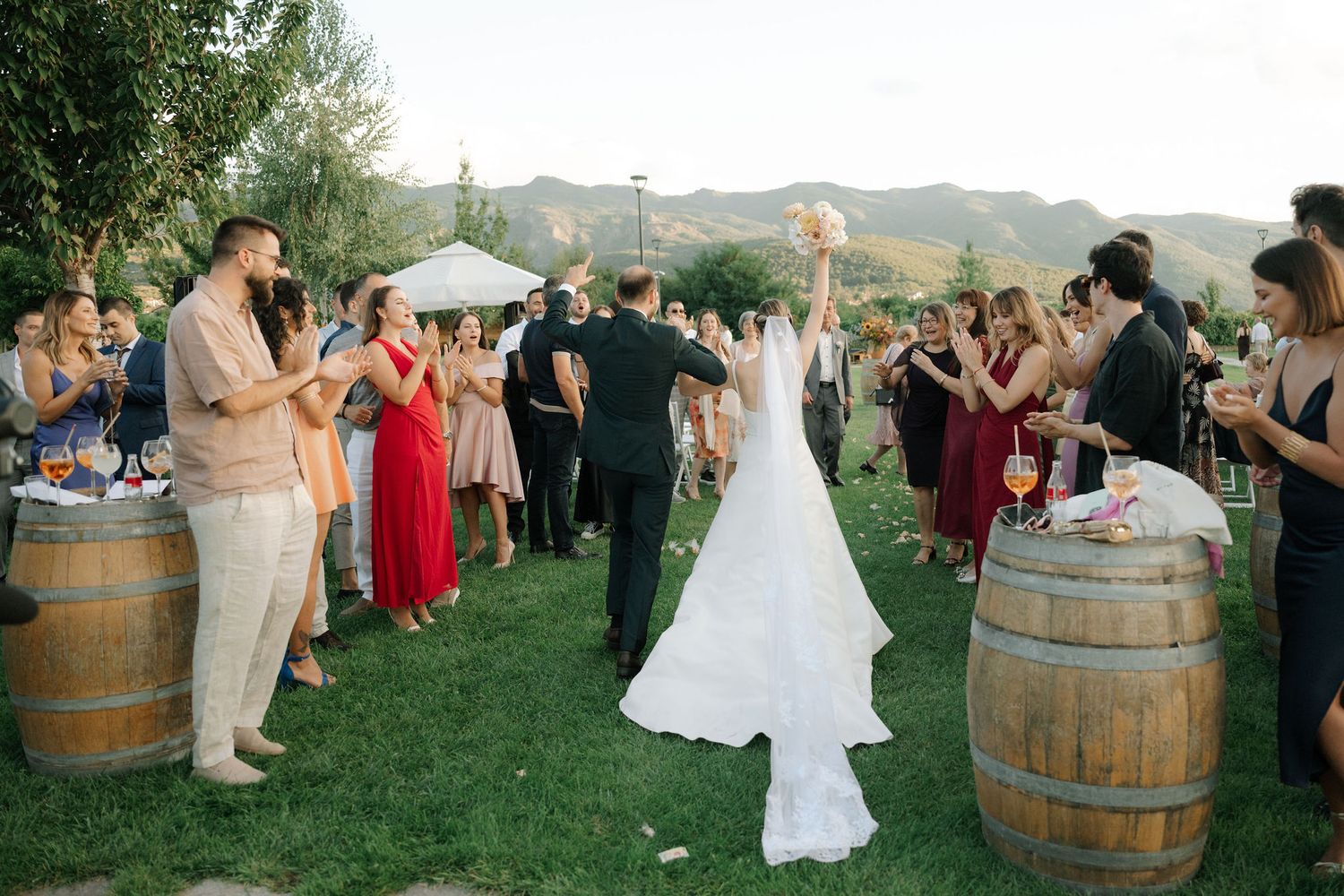 Newlyweds and wedding guests dance outdoors at a rustic vineyard reception with wooden barrels and string lights.
