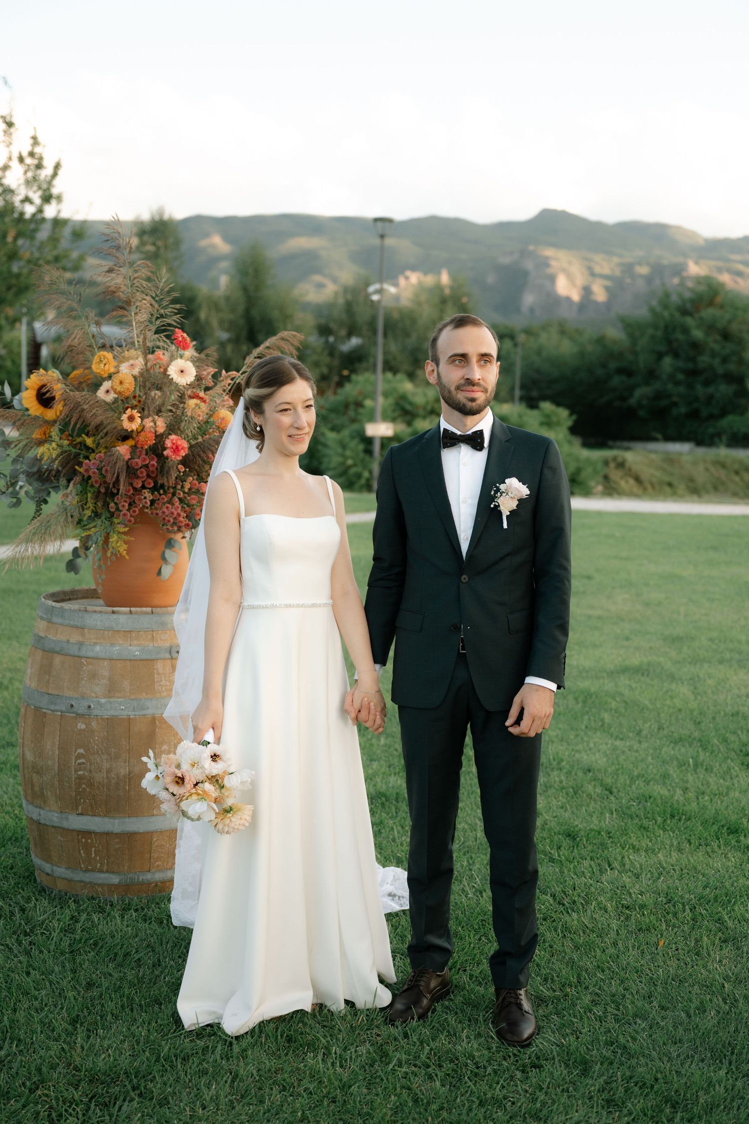 Couple in formal wedding attire poses in front of rustic wine barrel with autumn floral arrangements on grassy lawn.