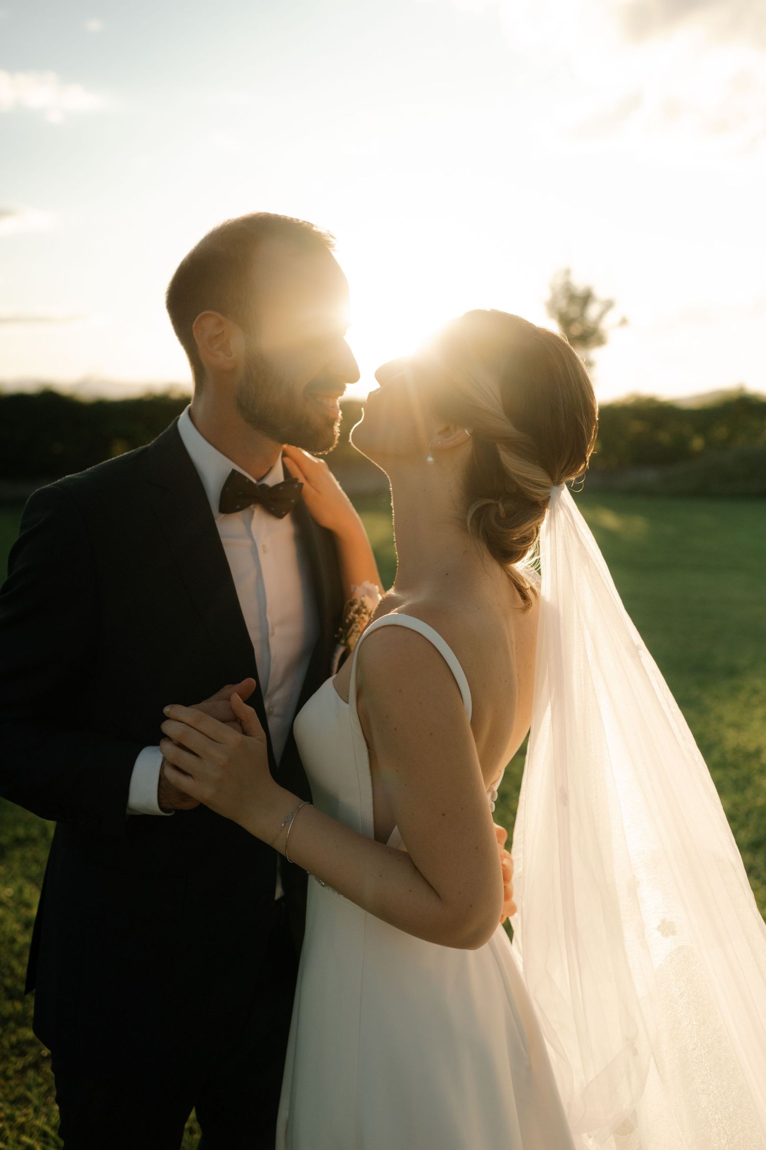 A romantic wedding couple embraces in a golden sunset on a grassy field, creating a silhouetted moment against the glowing sky.