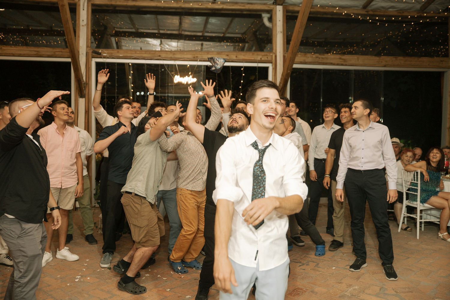 Festive wedding party dancing in formation under wooden beams and string lights at a rustic venue celebration.