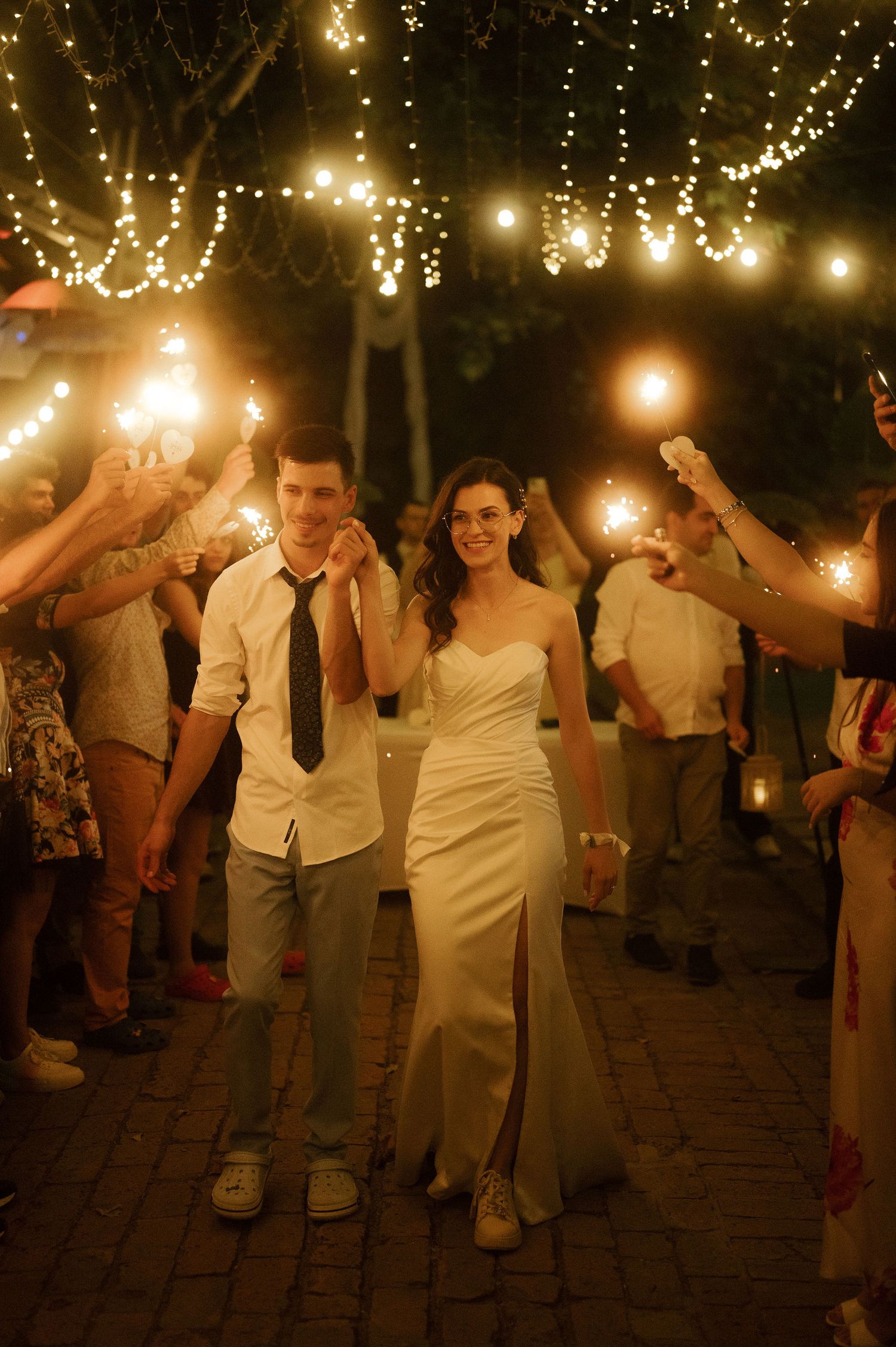 Wedding guests dance and celebrate on a brick patio under glowing string lights at an evening reception.