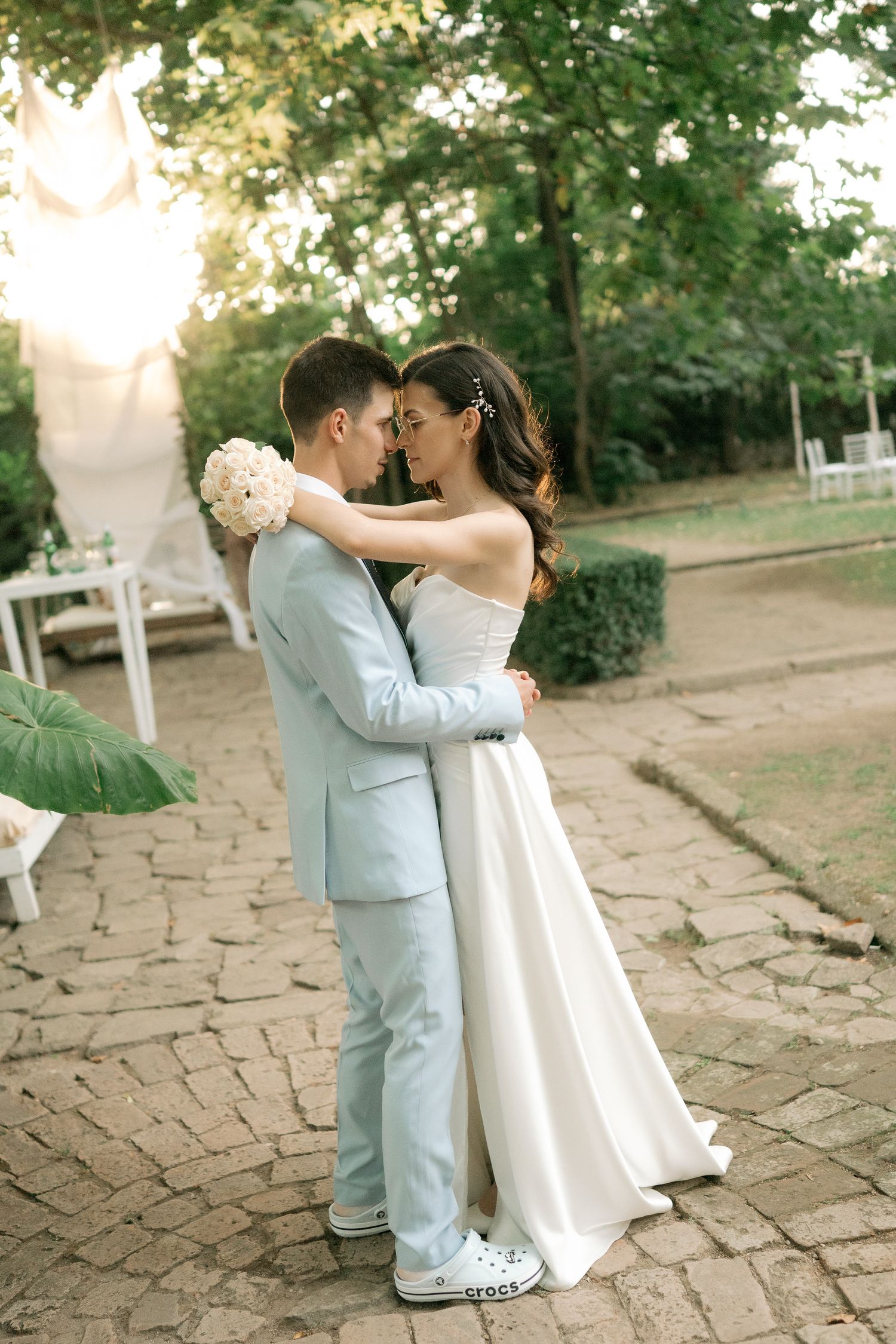 A romantic wedding couple embraces on a stone patio during golden hour, with trees and sunlight in the background.