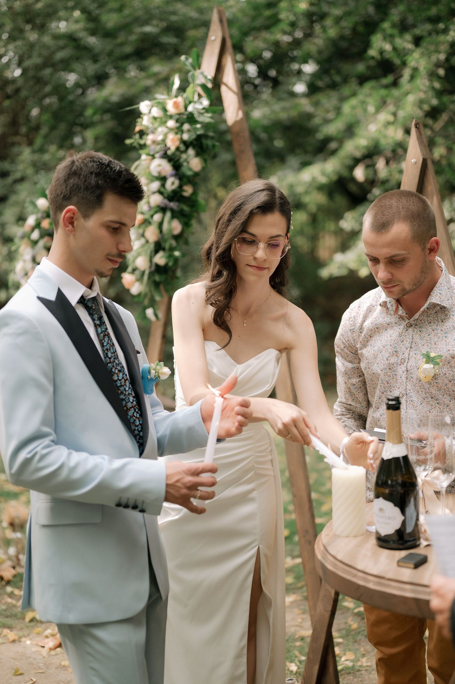 A couple shares a romantic moment while enjoying drinks at an outdoor wedding reception in a wooded setting.