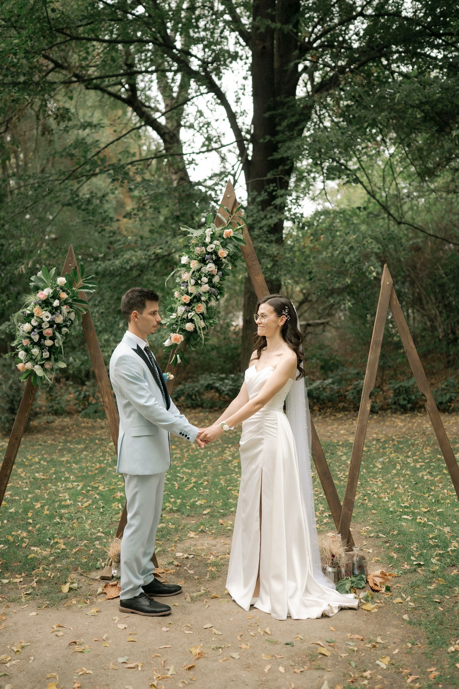 A couple shares a romantic moment during their outdoor autumn wedding ceremony surrounded by fallen leaves and tall trees.