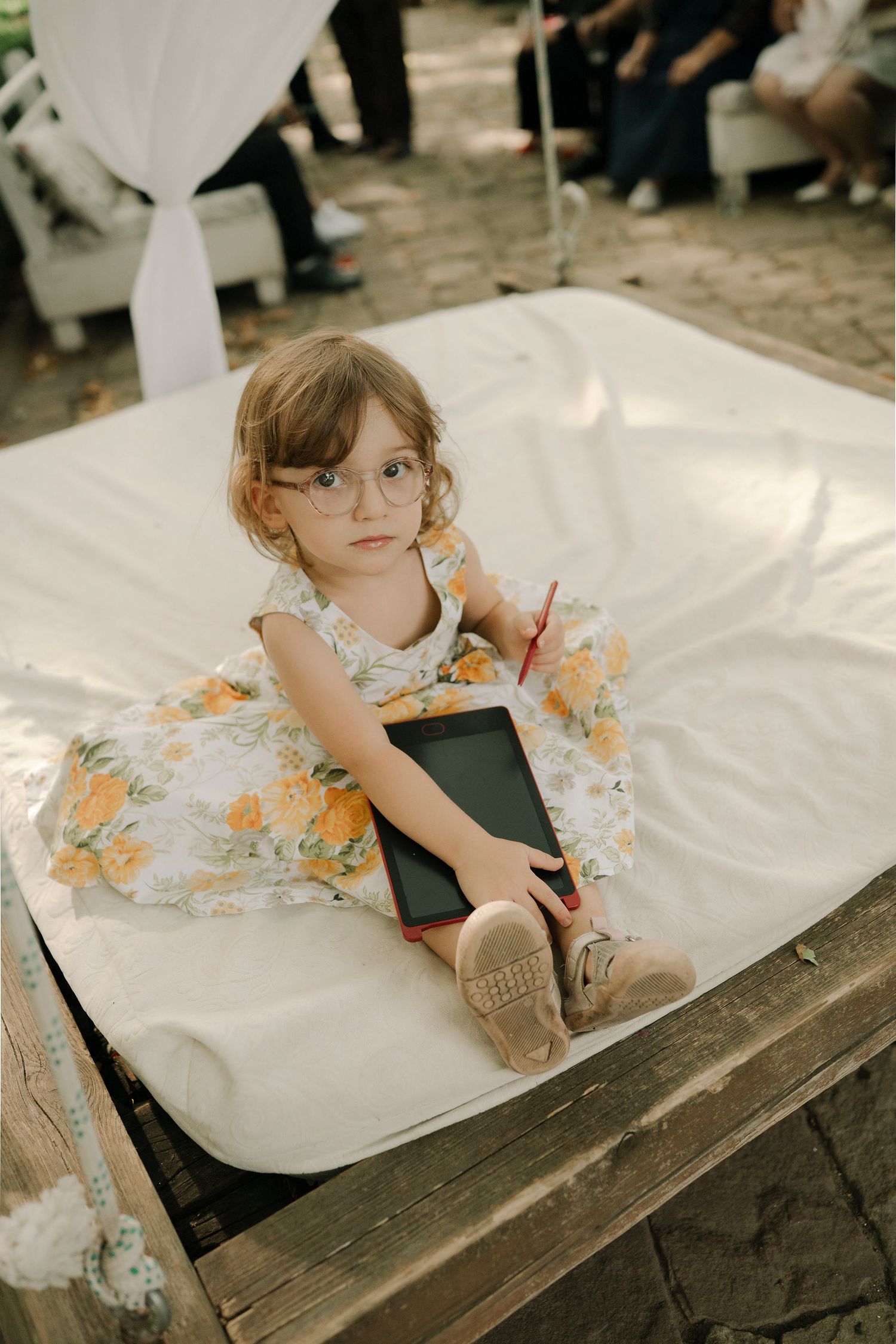 Young child in floral dress and glasses sits on white fabric outdoors while holding a book.