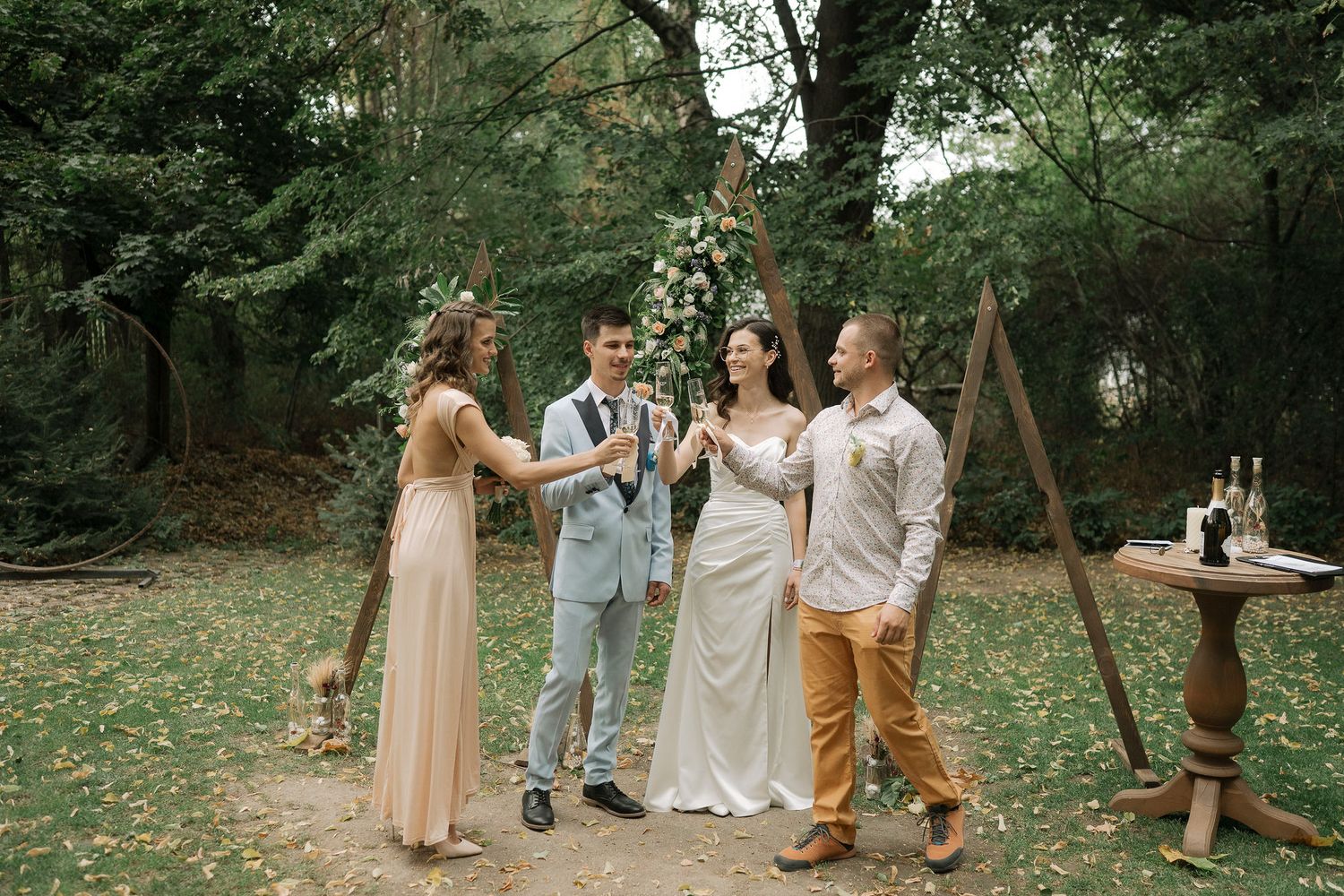 A group gathers for a casual outdoor celebration in a wooded setting with scattered autumn leaves on the ground.