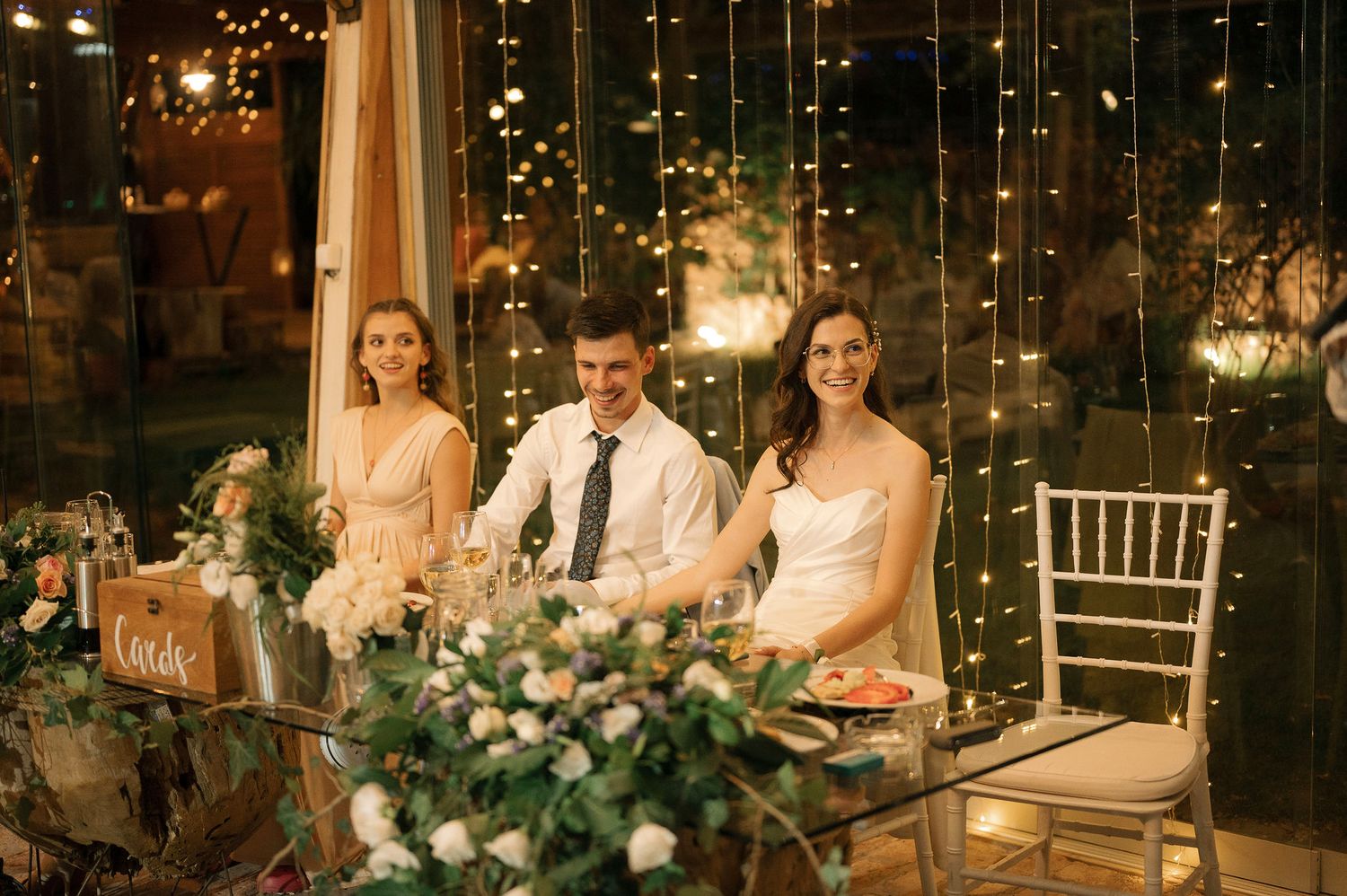 Romantic wedding reception scene with string lights, floral arrangements, and elegant table settings in a dimly lit venue.