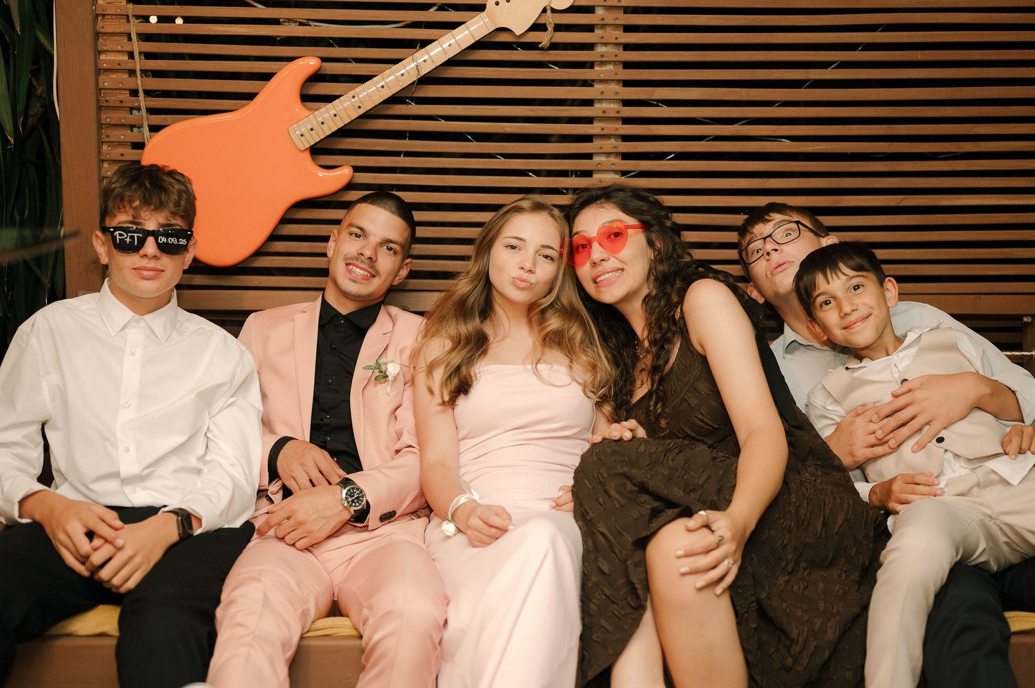 Group of friends in formal attire posing together on a couch at a party with retro-style lighting and window blinds behind them.