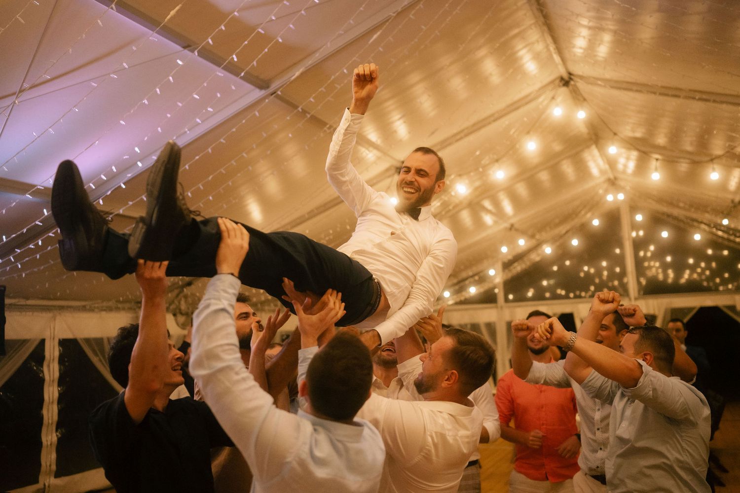 Wedding reception guests dance and celebrate under twinkling string lights in an elegant tent venue.