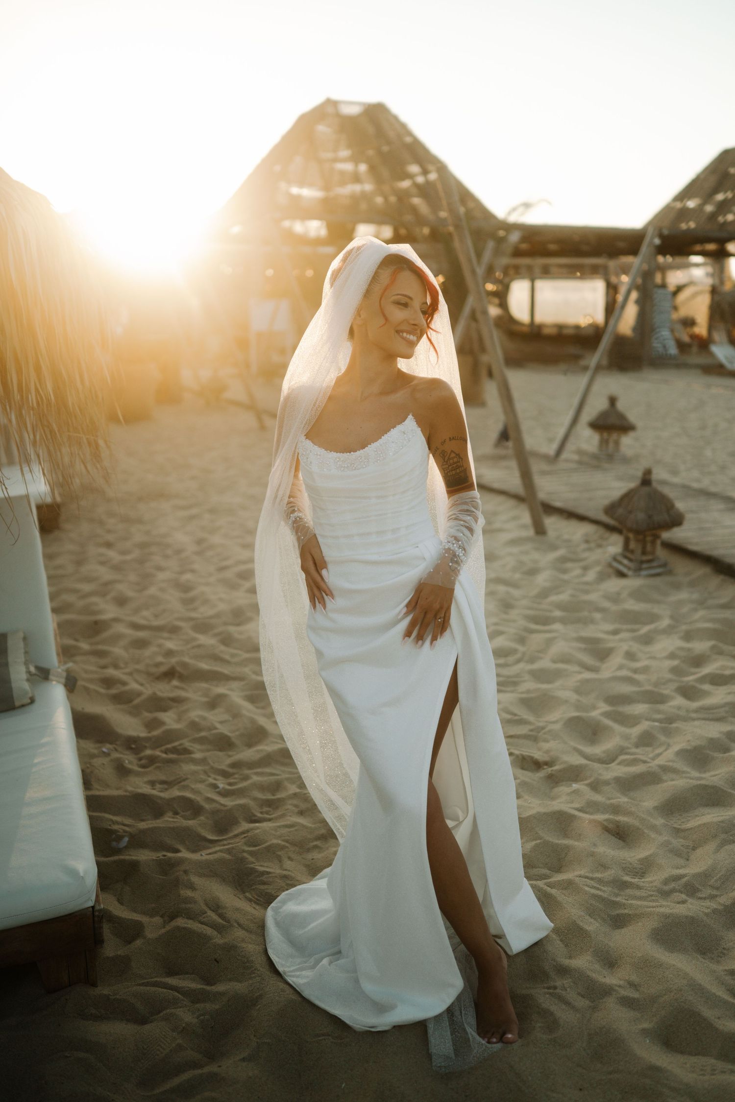 A bride in a white dress poses on a sandy beach at sunset with sunlight creating a warm golden glow.