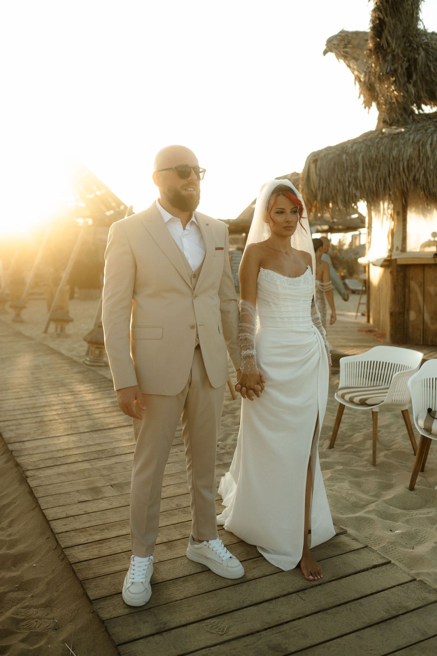 A couple walks hand in hand on a wooden boardwalk at sunset in elegant white and beige wedding attire.