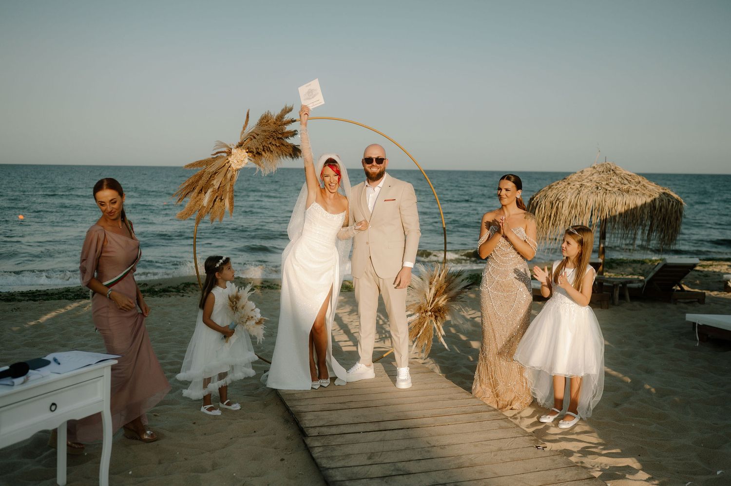 A wedding party poses on a wooden pier at sunset with beach umbrellas and the ocean in the background.