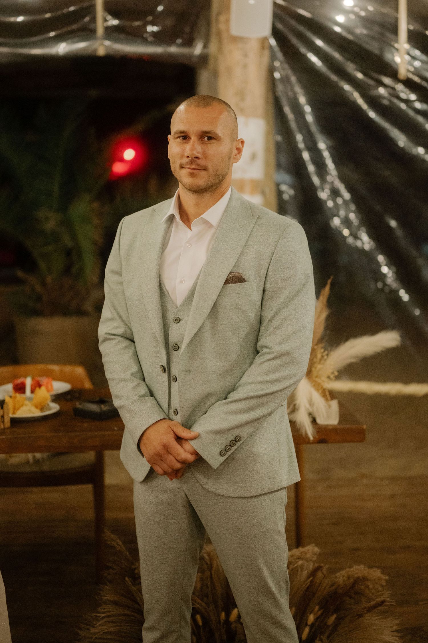 A groom in a light gray suit and white shirt poses at an outdoor wedding reception under string lights.