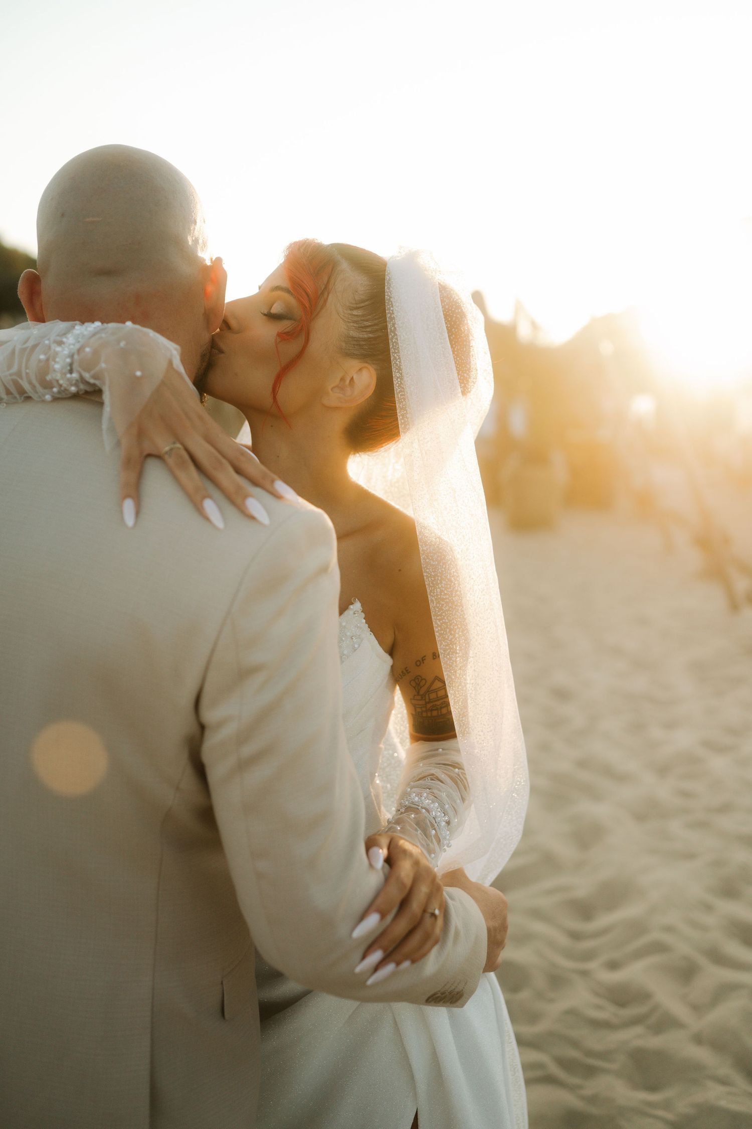 Bride in flowing veil embraces groom during sunset beach photoshoot with warm golden light filtering through.