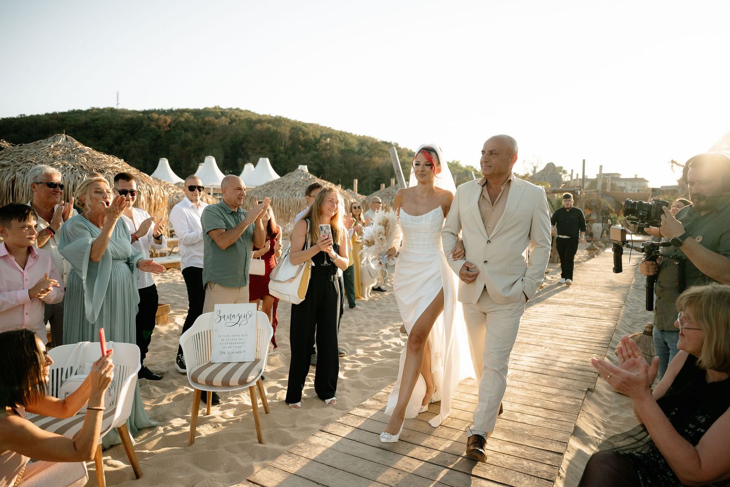 Wedding guests walk along a wooden boardwalk by the beach during sunset, creating a joyful processional line.