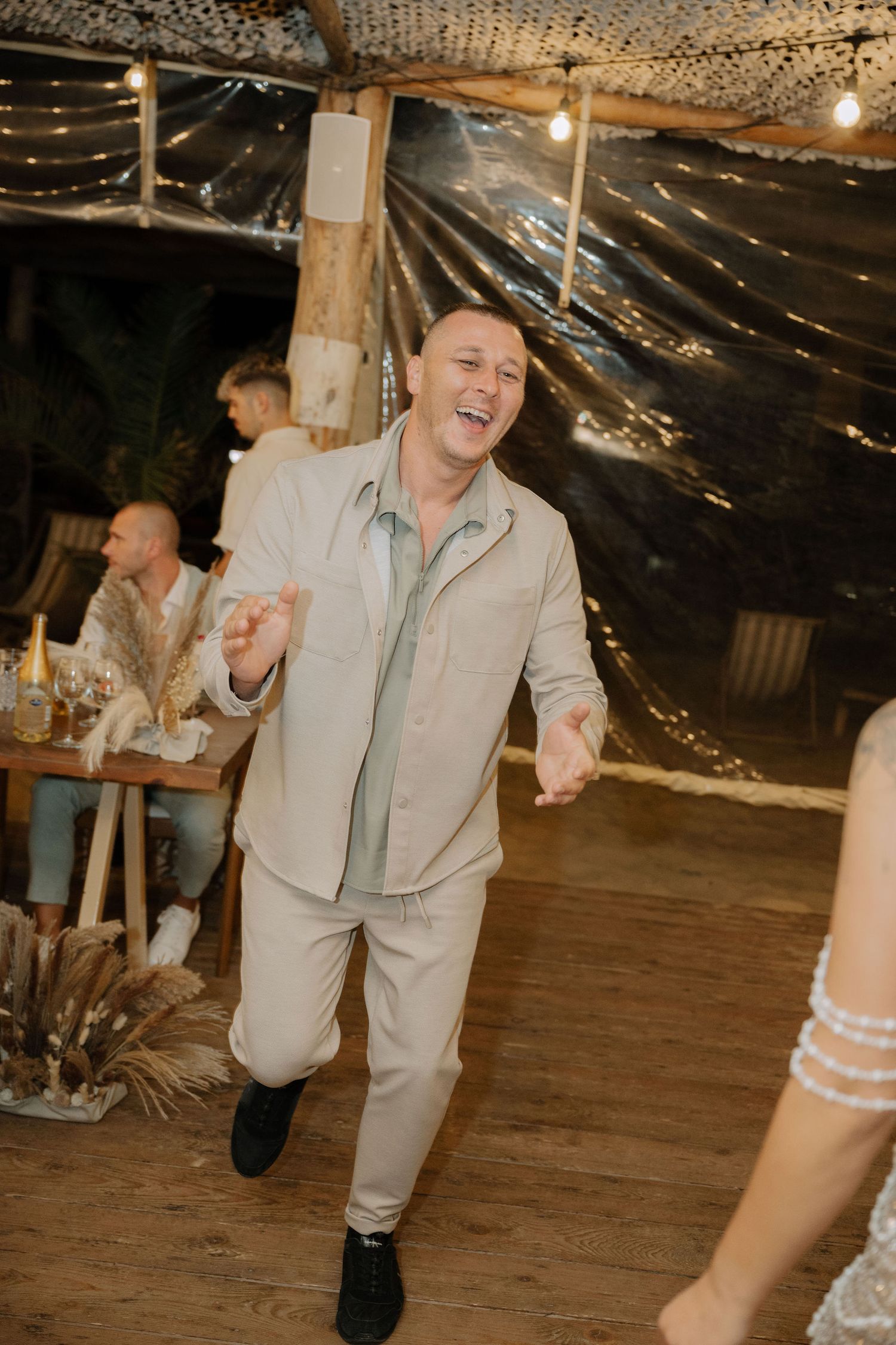 A person in a light colored suit dances energetically on a rustic dance floor under string lights.