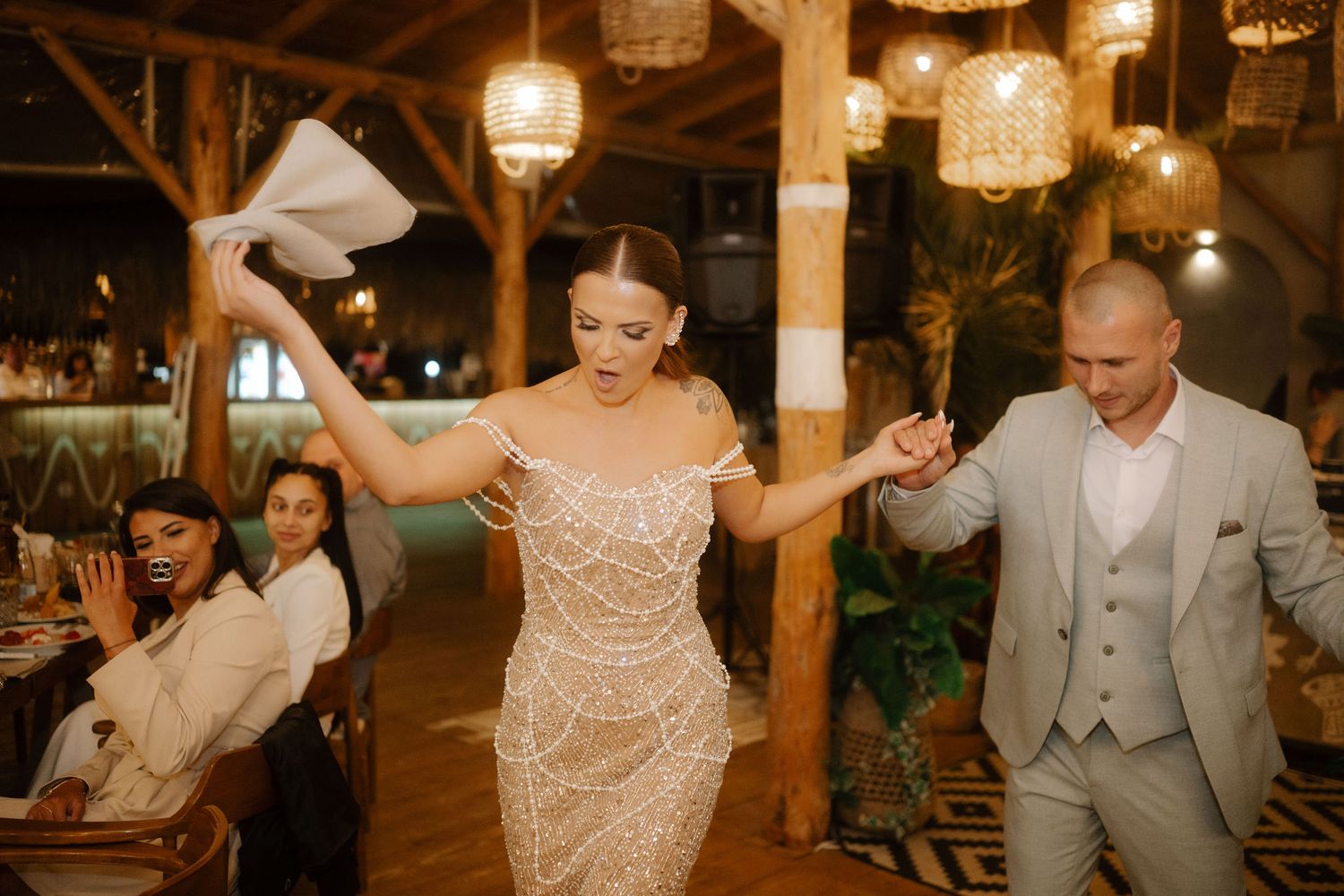 Wedding guests dance and celebrate on a rustic wooden dance floor under warm glowing pendant lights at an evening reception.
