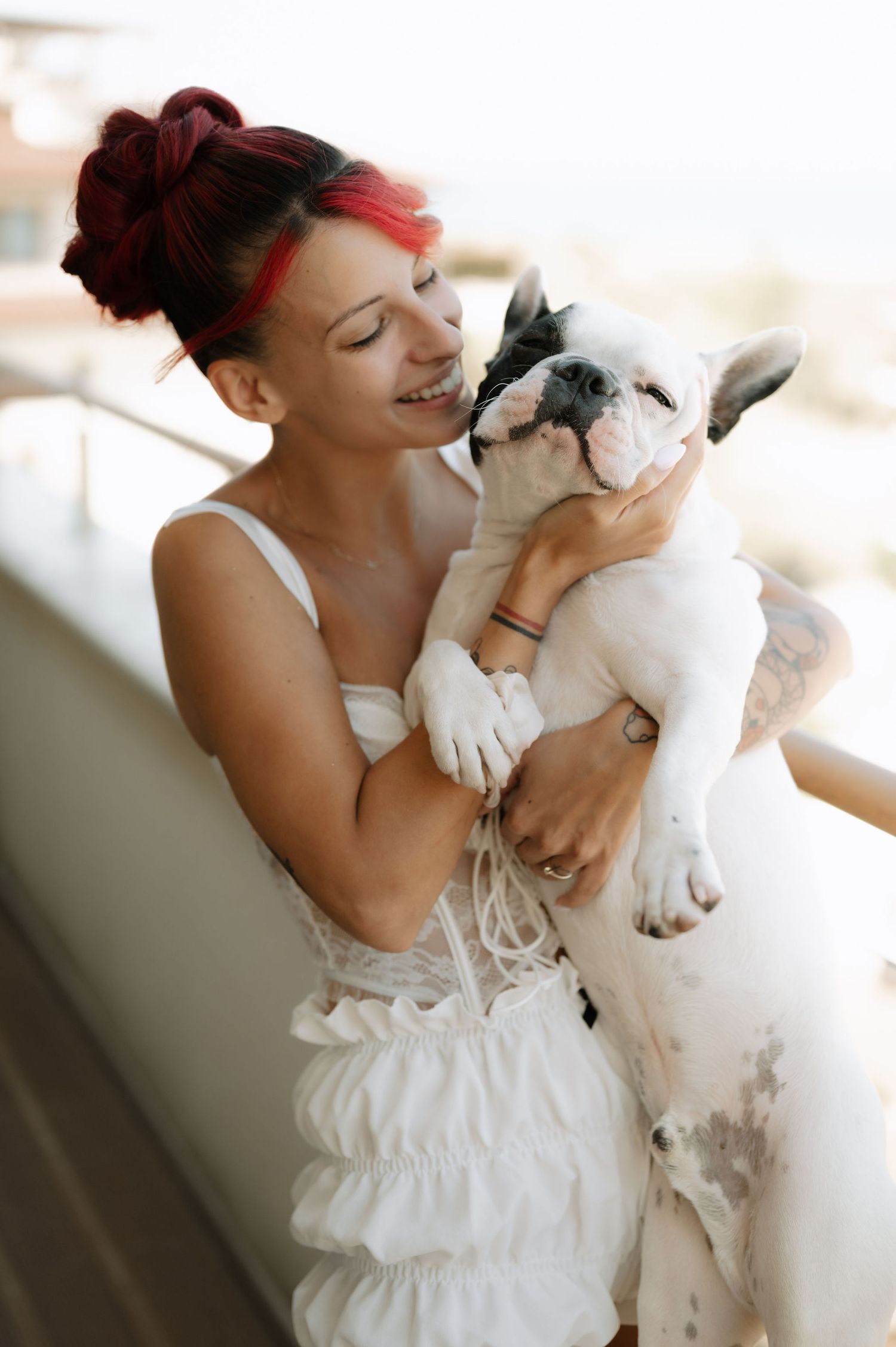 Woman in white dress shares tender moments with small white dog on couch in natural light setting.
