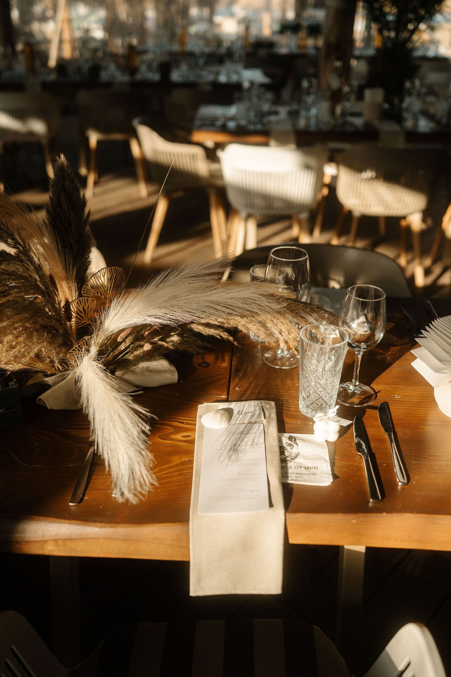 Elegant table setting with pampas grass decor, wine glasses and cutlery in warm sunlight at a restaurant.