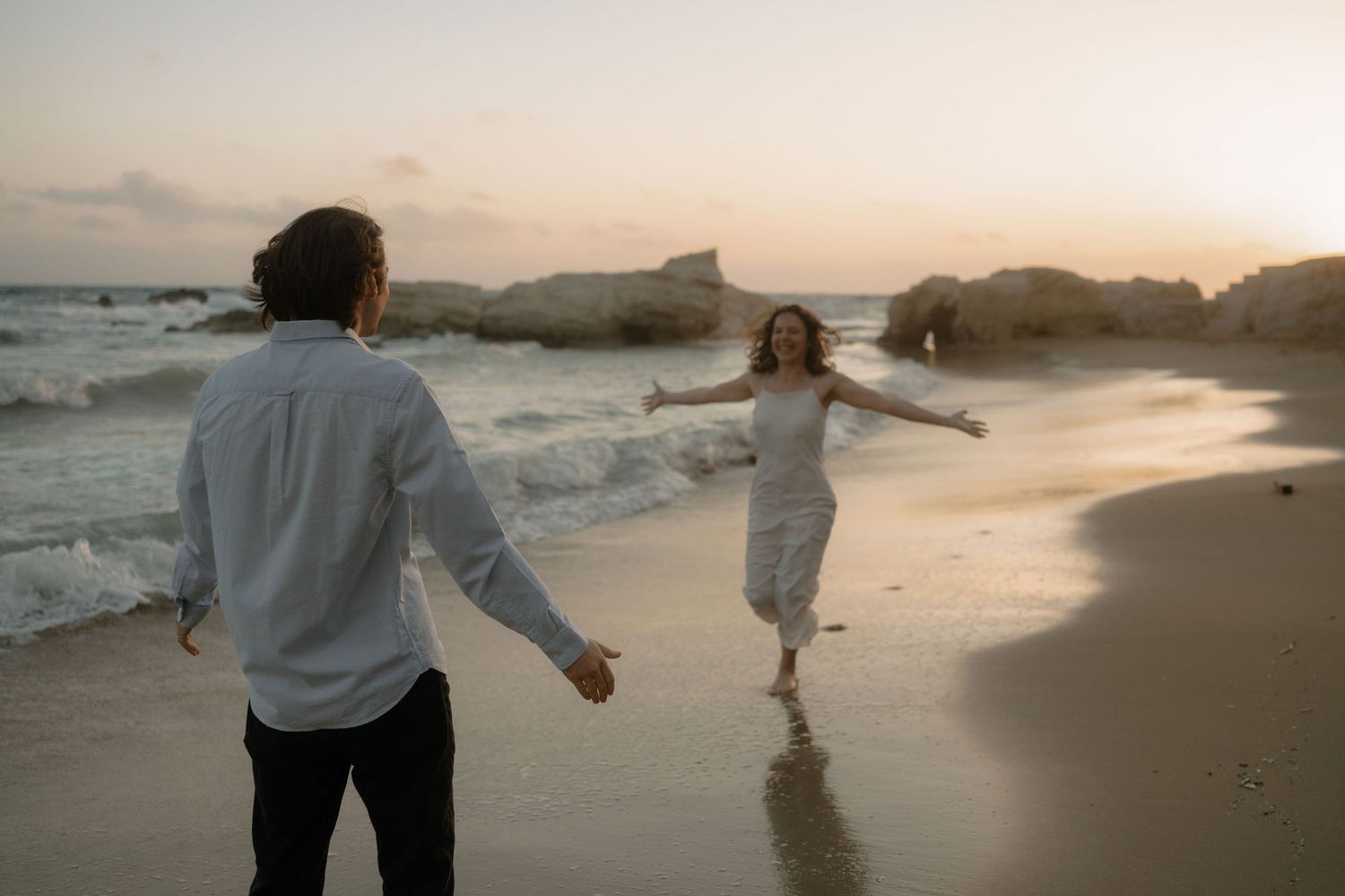 Couple running on a sandy beach at sunset, woman in white dress with arms outstretched, waves crashing behind her.
