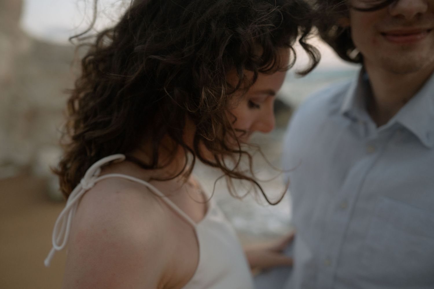 Romantic couple embracing outdoors, woman with curly hair in white dress, soft natural light photography.