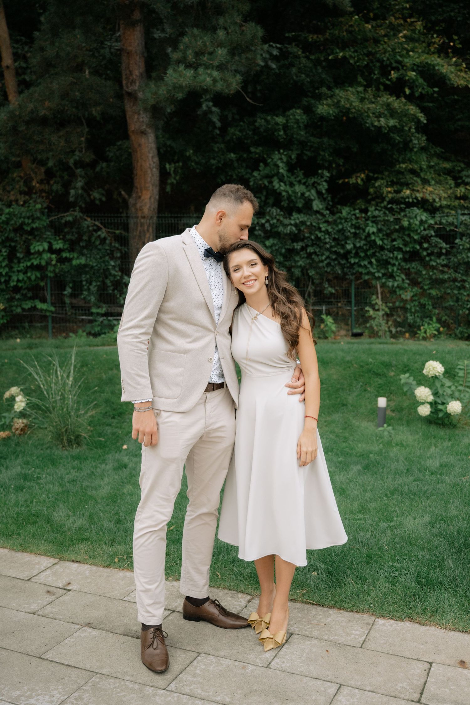 A couple in matching light beige outfits embrace on a stone patio surrounded by lush garden landscaping.
