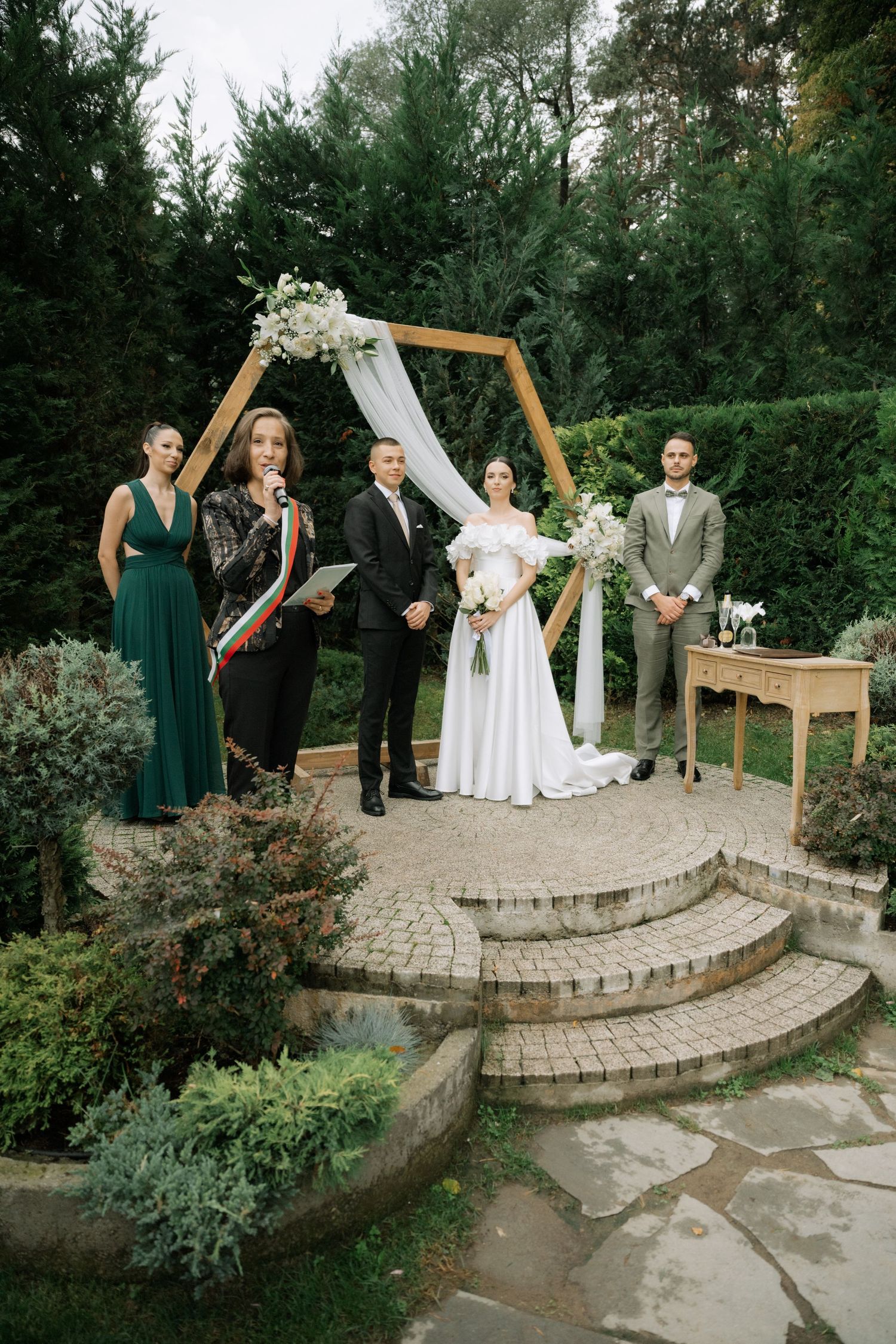 Wedding party gathers on curved stone steps in a garden setting during outdoor ceremony.