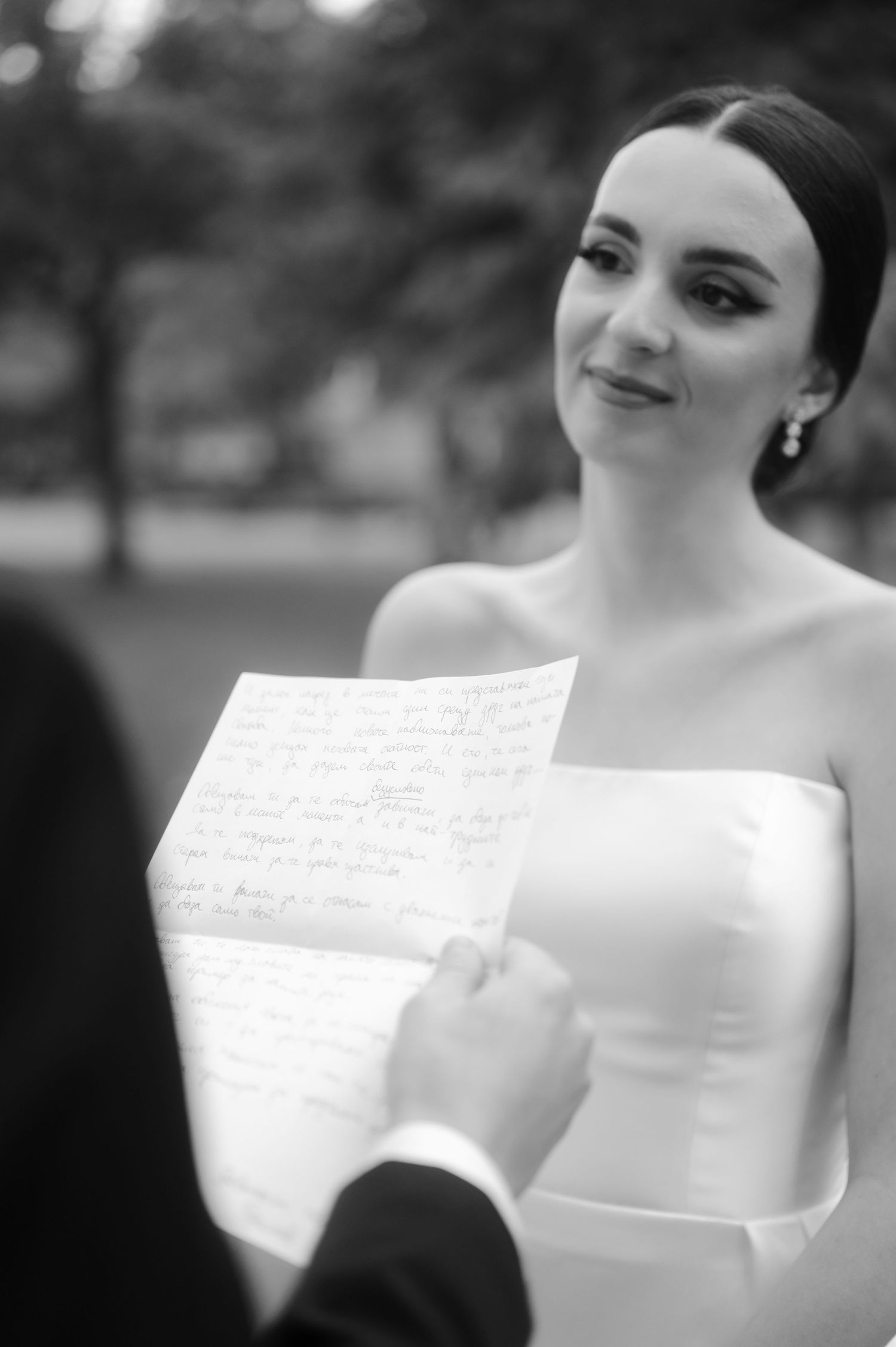 Black and white photo of a bride in a strapless gown reading from a paper during an outdoor wedding ceremony.