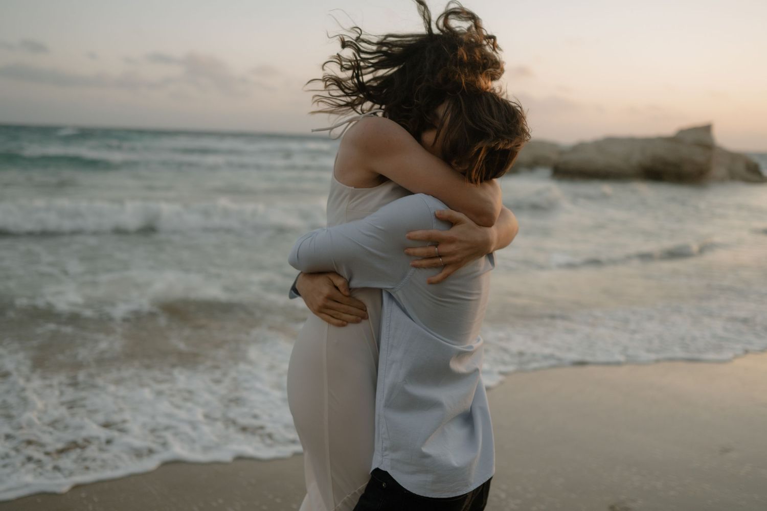 Couple embracing romantically on a sandy beach at sunset with ocean waves in the background.