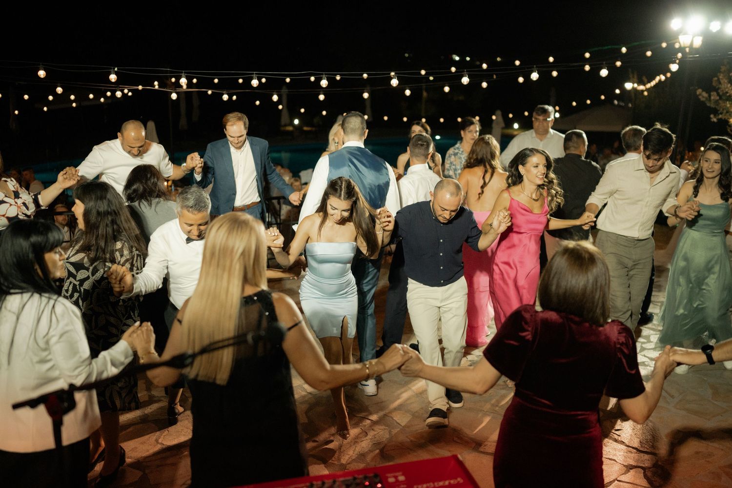 People dancing together in a line at a festive outdoor event with string lights overhead.