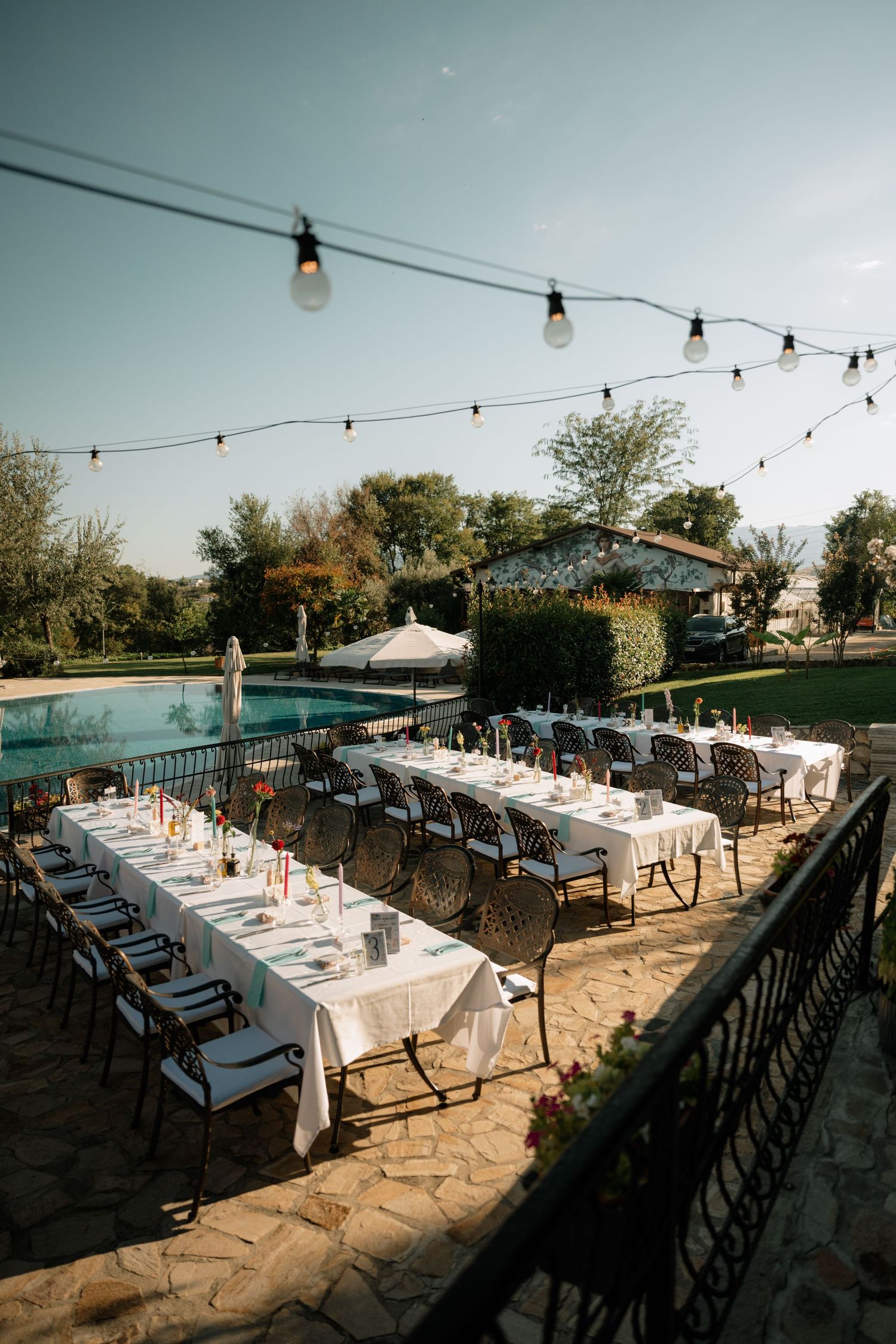 Outdoor dining setup with string lights and white tablecloths overlooking a swimming pool at sunset.