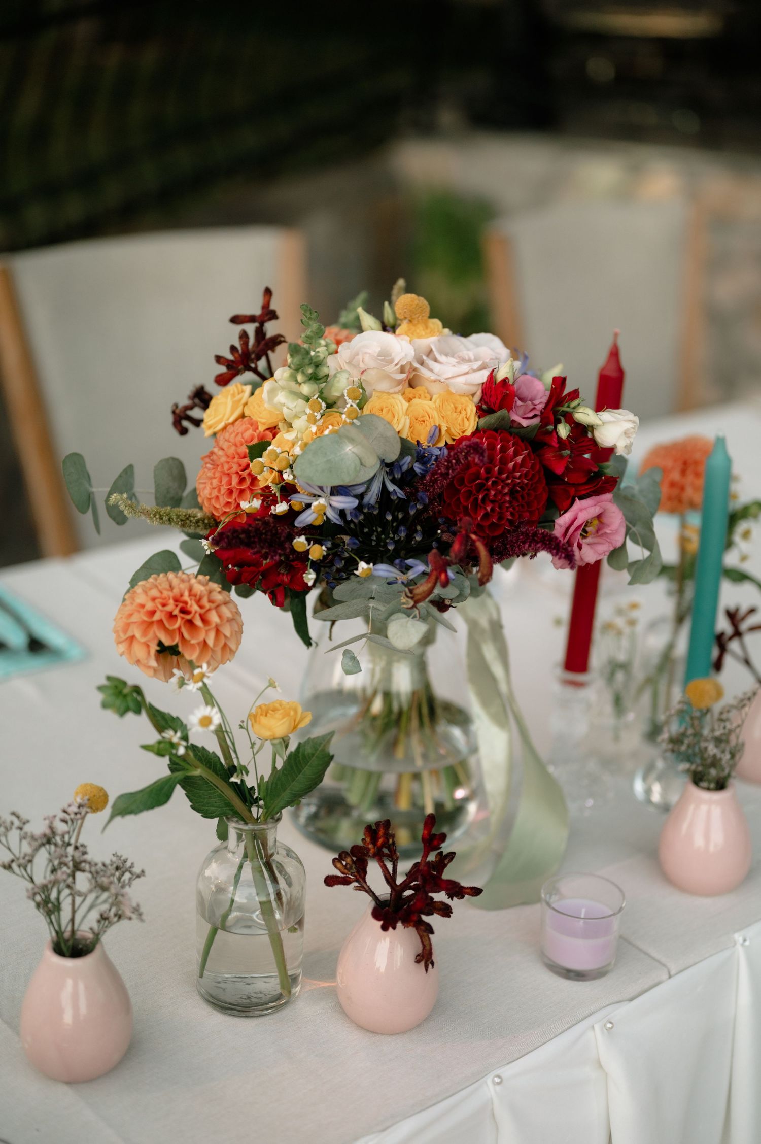 Colorful floral centerpieces in pink vases and clear jars line a white tablecloth at a wedding reception.