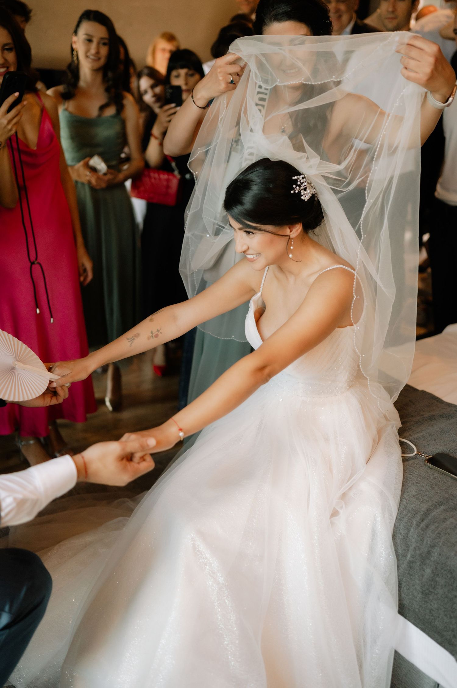 A bride in a flowing white wedding dress and veil dances joyfully at a reception surrounded by guests.