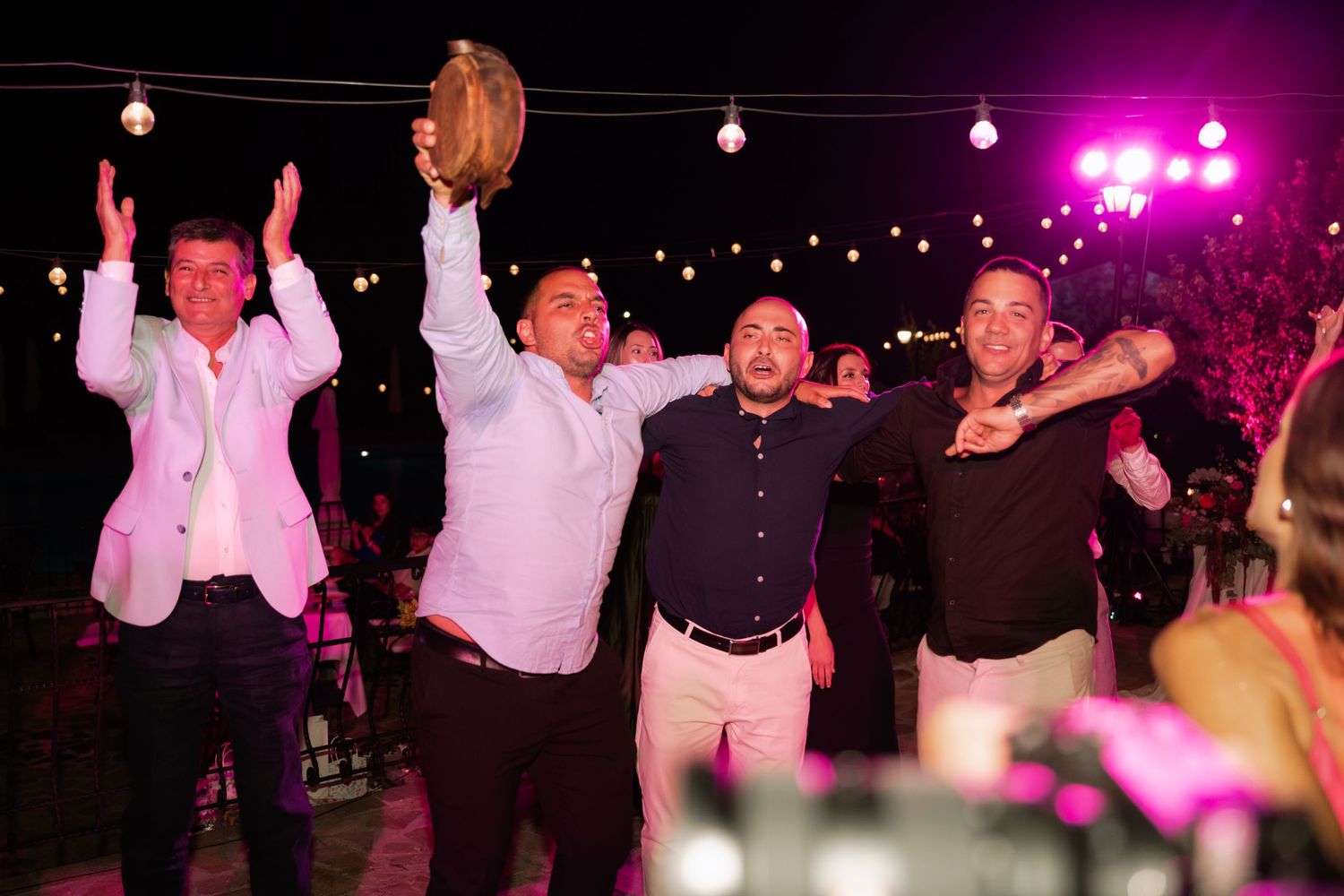 People dancing and celebrating at a lively wedding reception with purple lighting and a festive atmosphere.