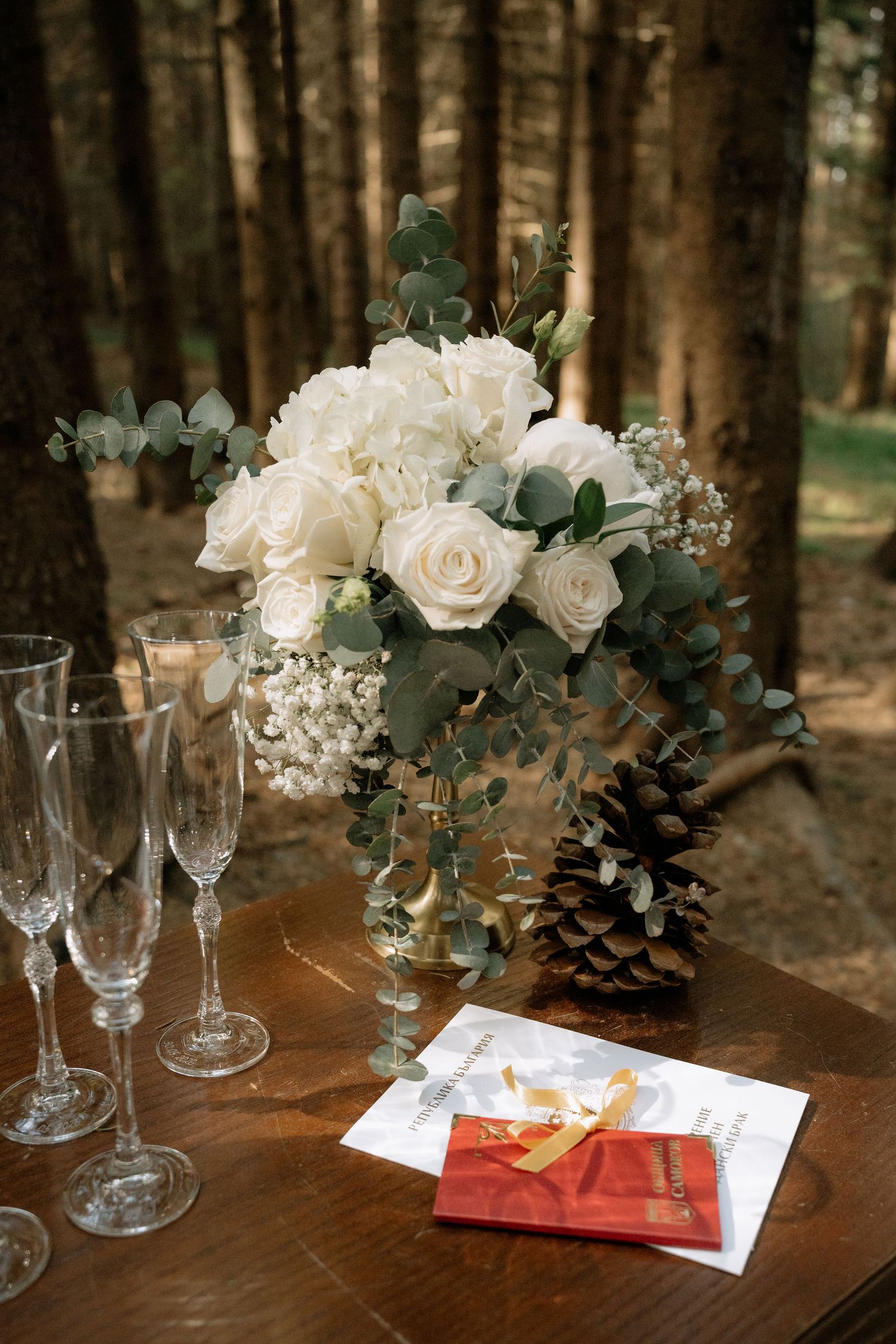 Elegant wedding table with white roses, eucalyptus bouquet, champagne flutes, pine cone, and red gift box in forest setting.