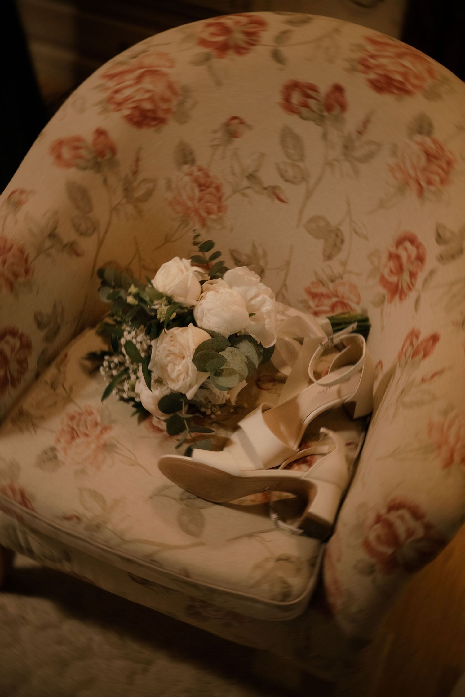 Bridal heels and white floral bouquet resting on a vintage floral armchair for a romantic wedding detail shot.