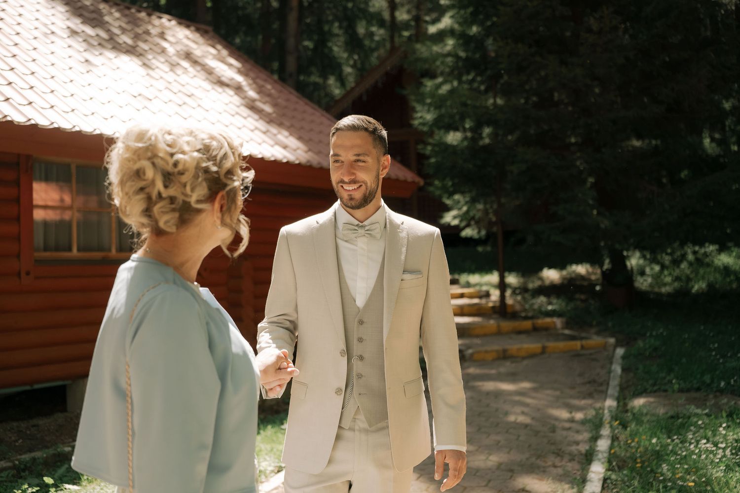 A smiling groom in a beige suit holds hands with a woman in a light blue dress outdoors near a wooden cabin.