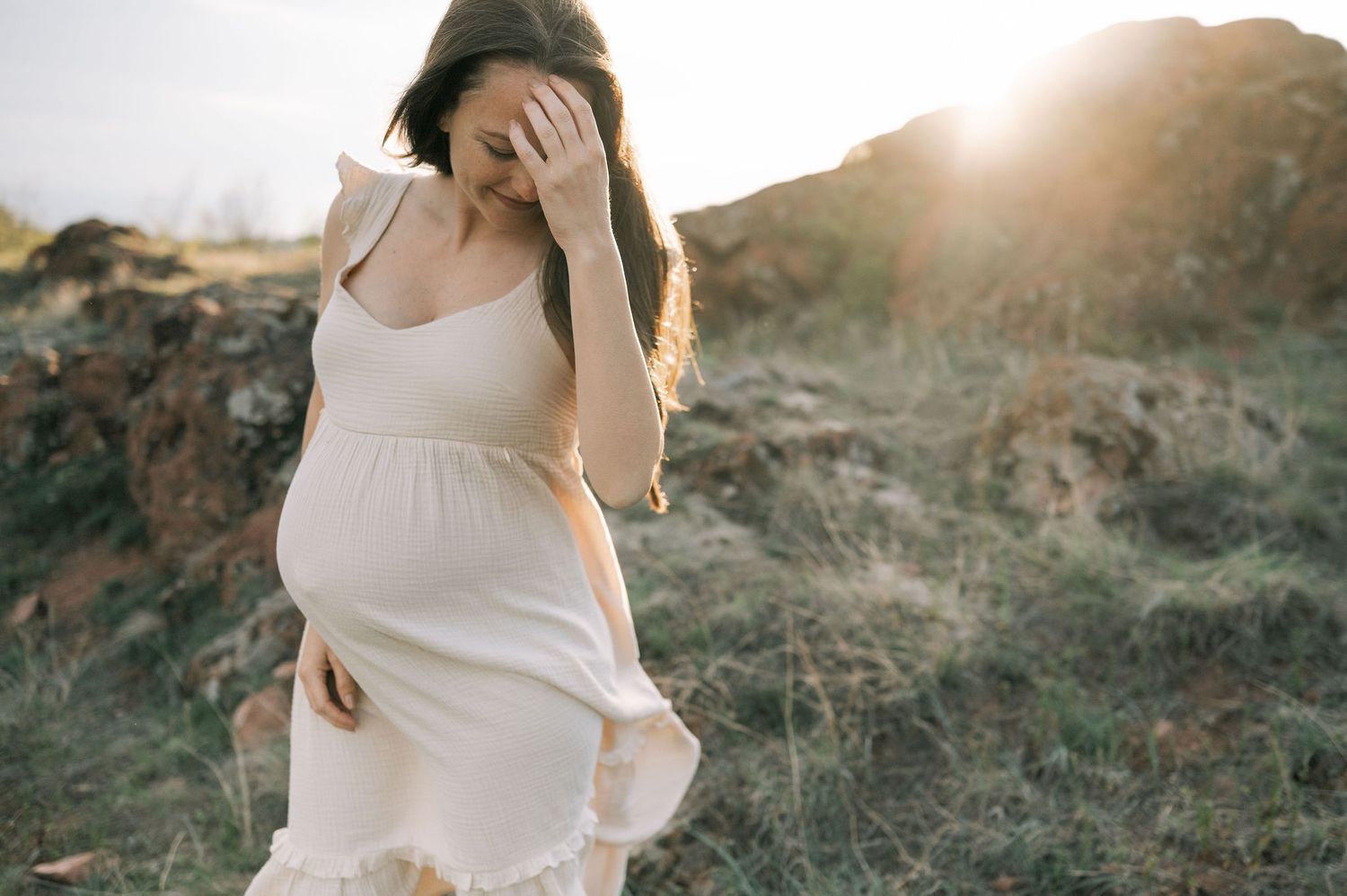 A pregnant woman in a flowing white dress stands on a rocky hillside at sunset with golden light behind her.