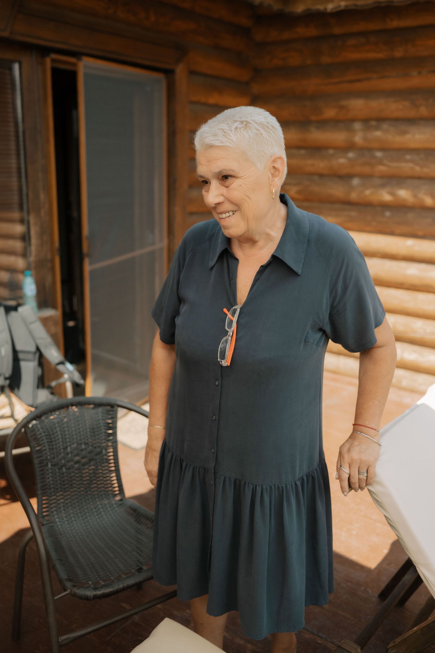 Person in a navy blue dress stands on the porch of a rustic log cabin on a sunny day.