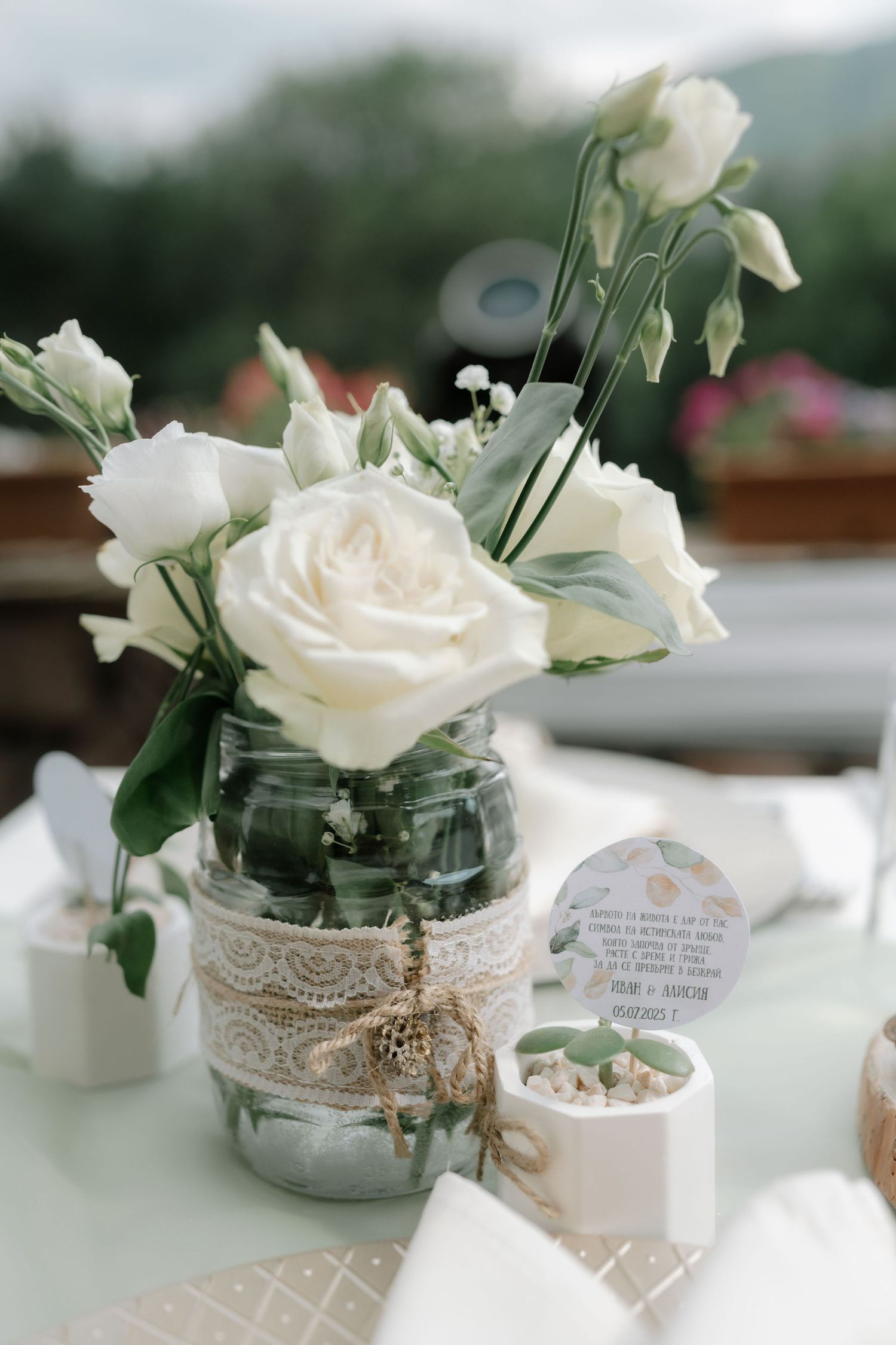 White roses in a mason jar decorated with lace and twine sit on a table with rustic wedding decor.