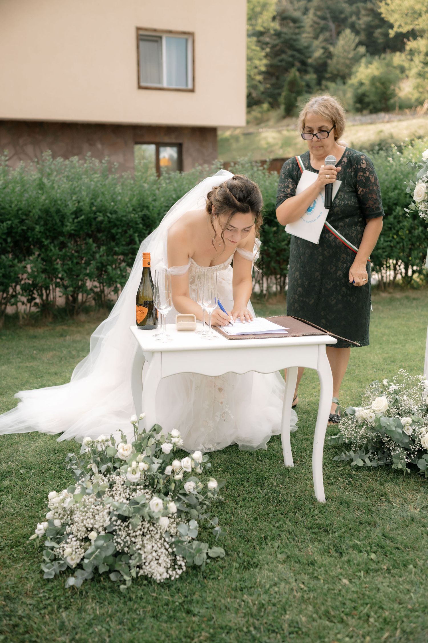 A wedding ceremony signing moment at an elegant white table outdoors with floral decorations on a green lawn.