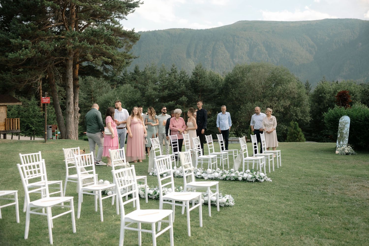 White wooden chairs arranged in rows on a lush green lawn with mountains and trees in the background for an outdoor ceremony.