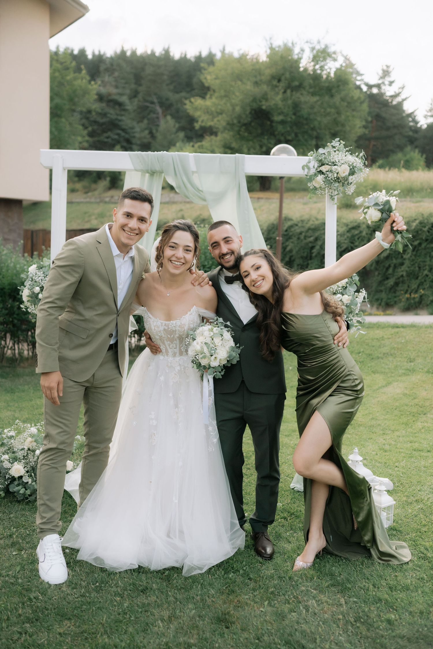 Wedding party members pose playfully on grass in coordinated olive green and black attire at outdoor celebration.