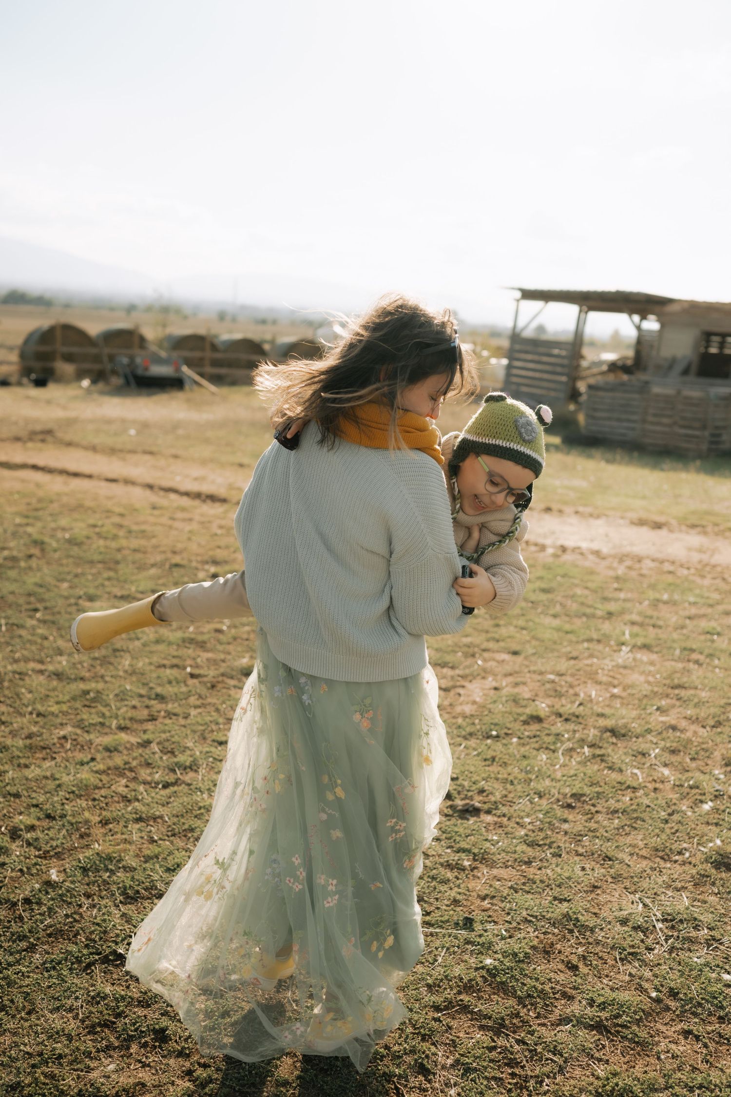 Two people in light-colored outfits dance and spin together in a rustic outdoor field at golden hour.