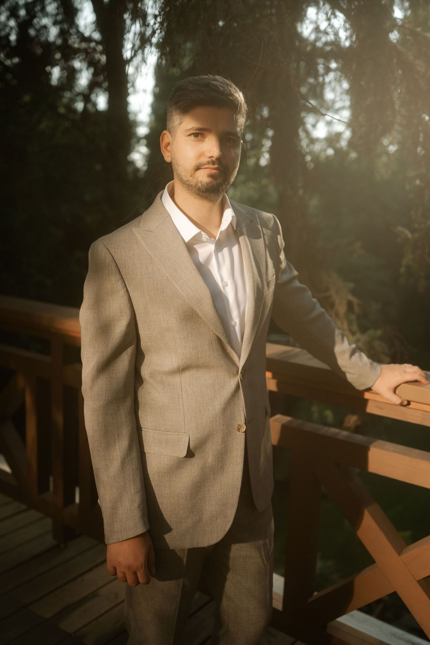 A person wearing a light gray suit and white dress shirt stands outdoors in warm sunlight near wooden benches.