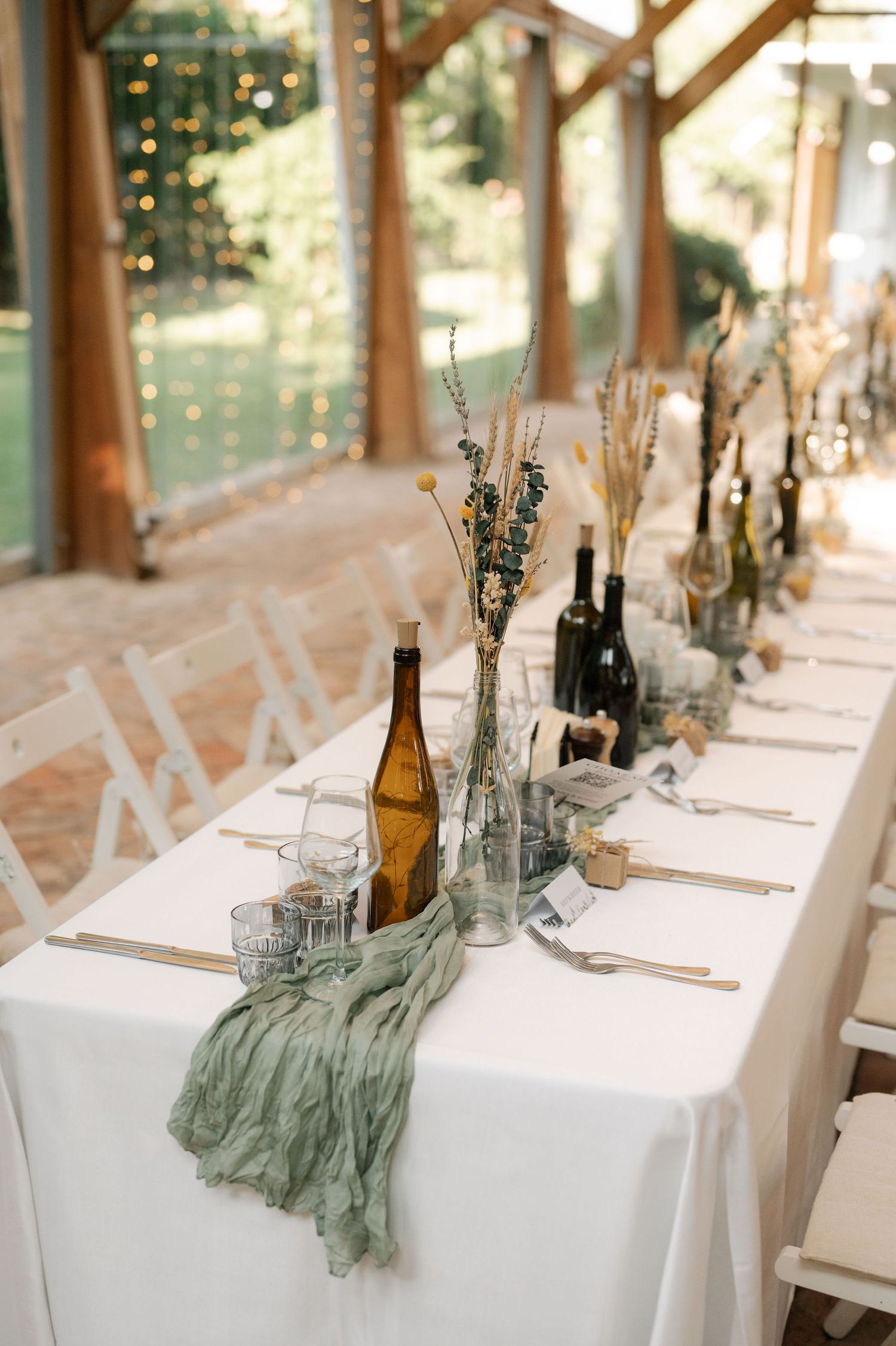 Elegant wedding table settings with white linens, sage green accents, and wine bottles in a rustic wooden venue.