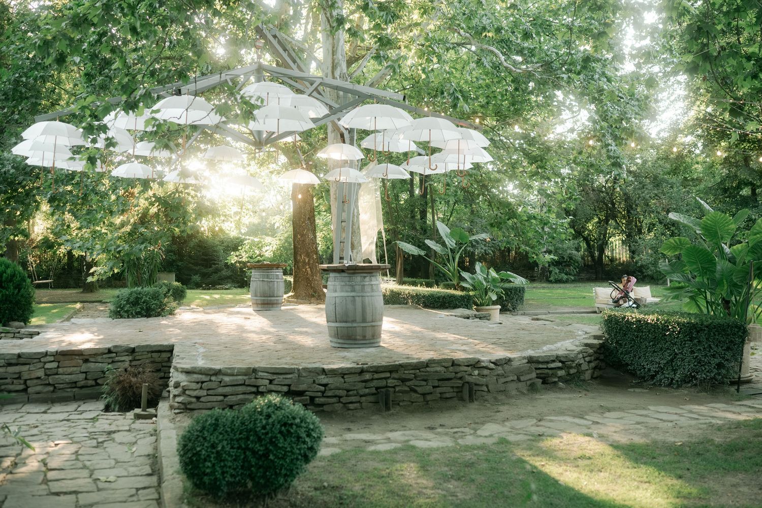 A serene garden pathway with stone walls and lush greenery leads to a sunlit clearing on a summer day.