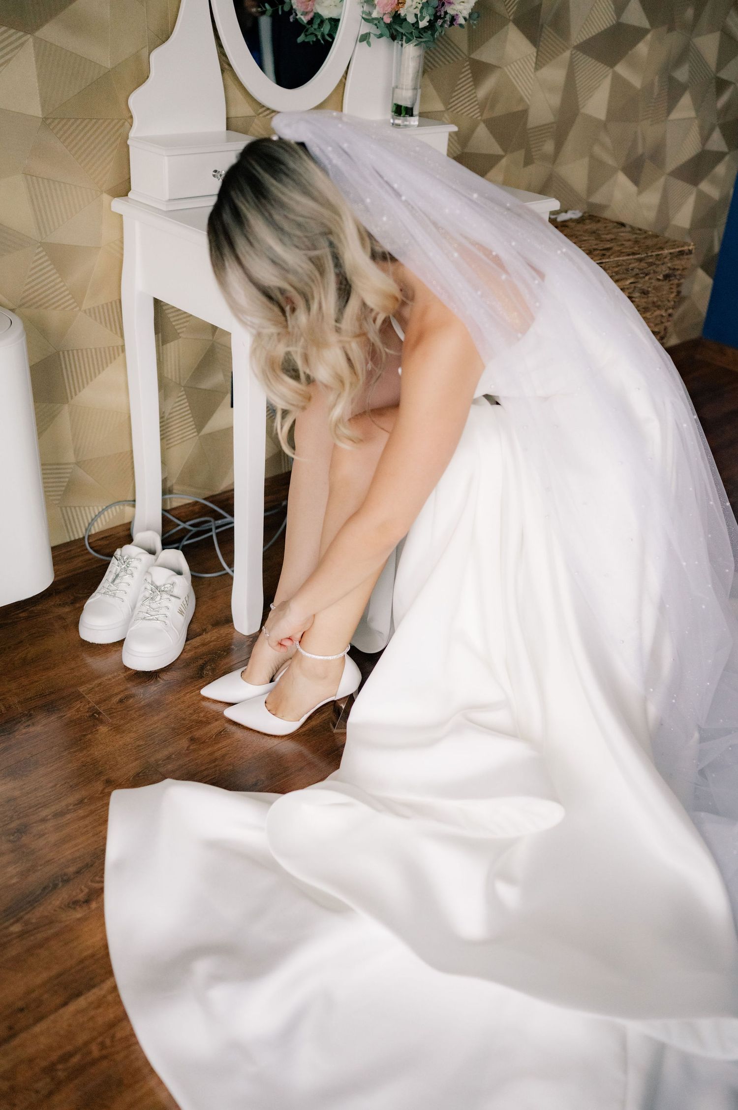 A bride in a flowing white wedding gown sits gracefully on wooden flooring while holding a bouquet.